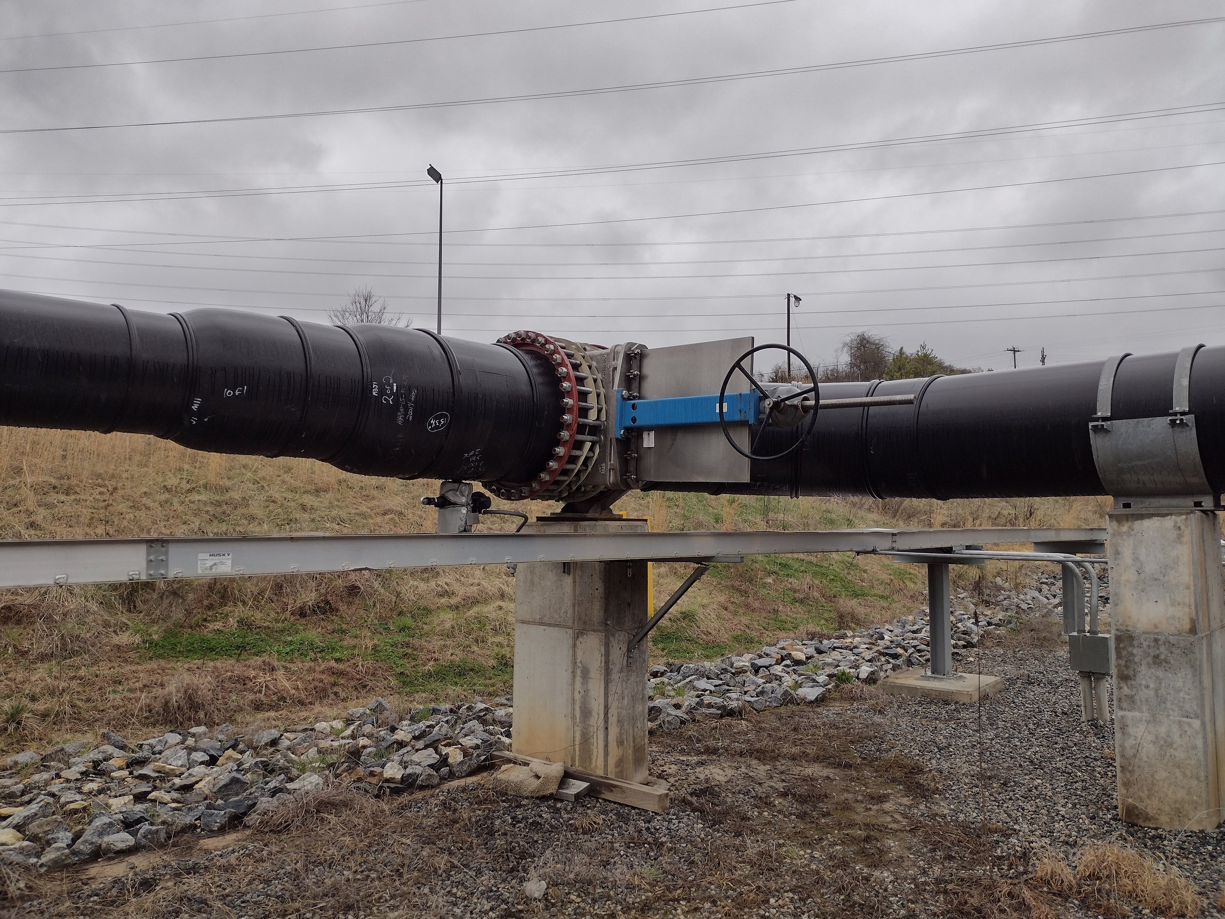 A large pipeline is supported by concrete and metal structures, running across a grassy area with rocks, under an overcast sky with power lines in the background.