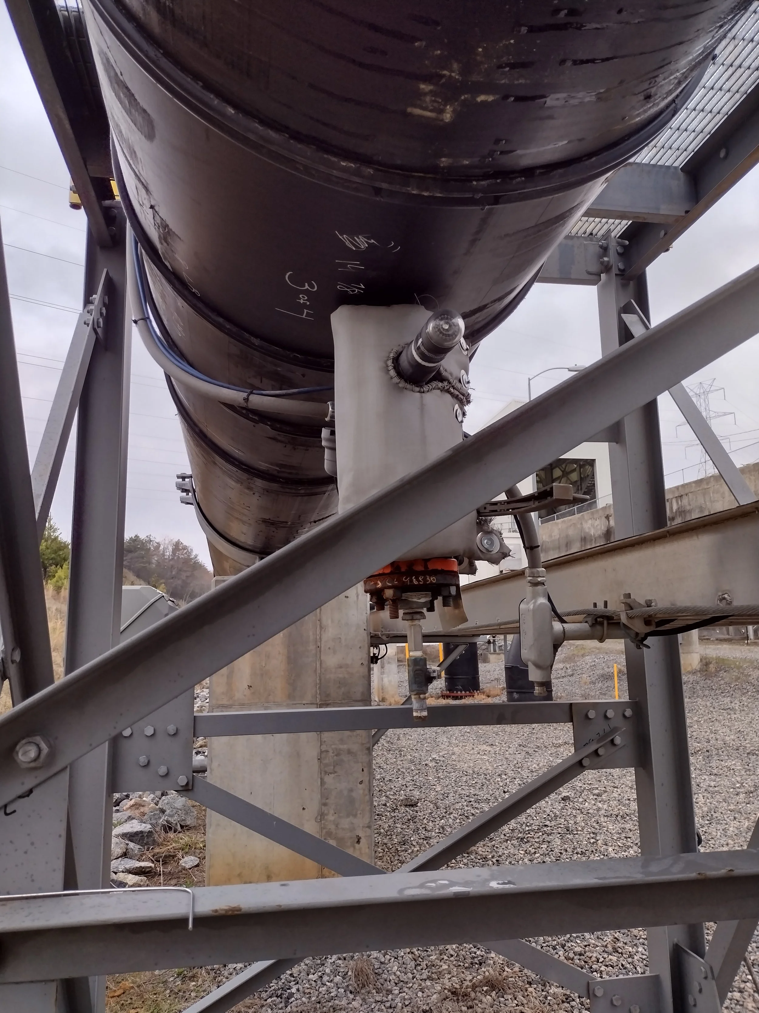 Large black pipeline elevated on a metal support structure outdoors. The pipeline has writing on it and is connected to a smaller pipe with a valve. The background shows a cloudy sky, hills, and power lines.