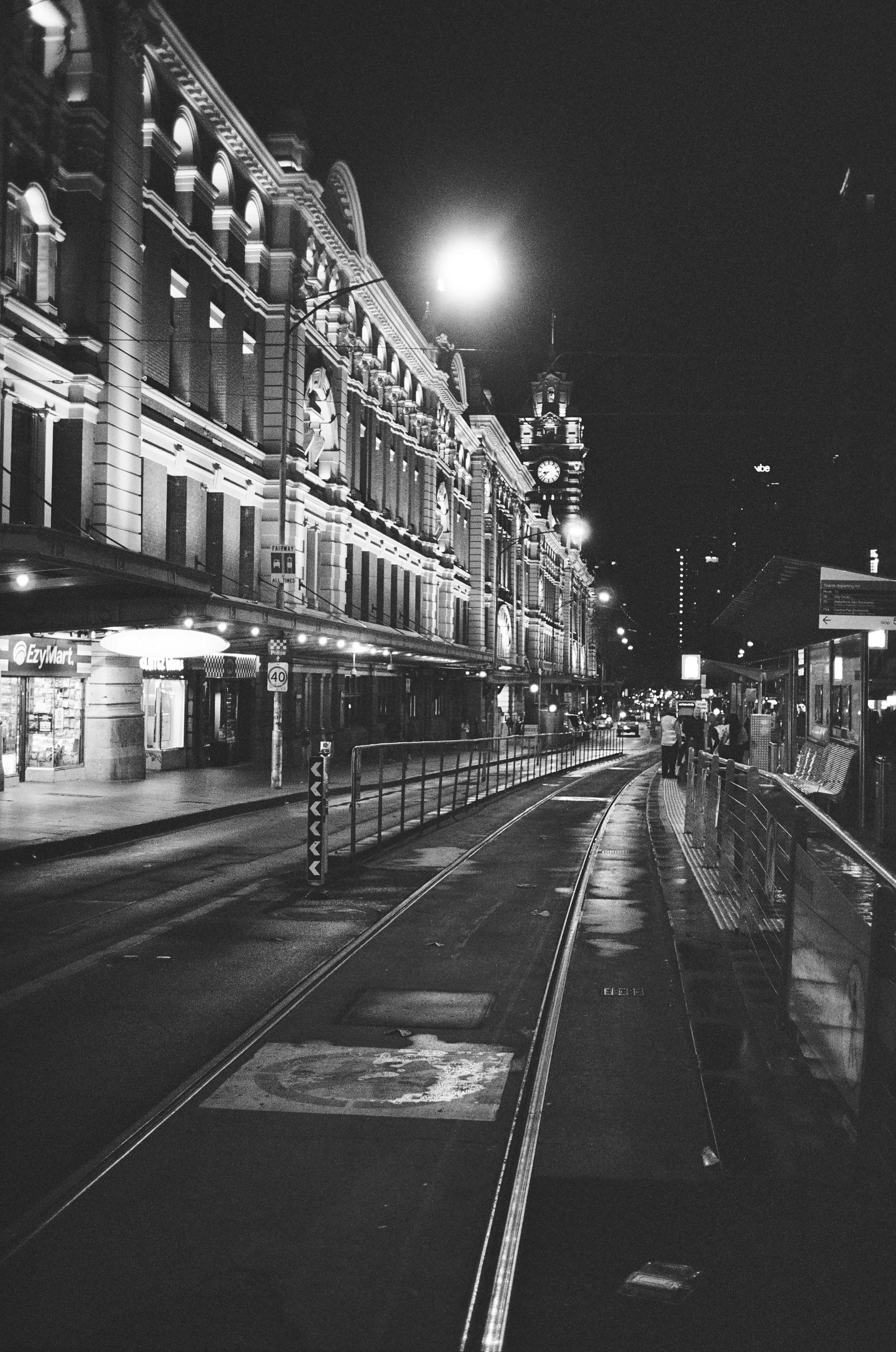 Nighttime scene of a city street with historic building facades, bright streetlights, a clock tower, tram tracks, and a few pedestrians.