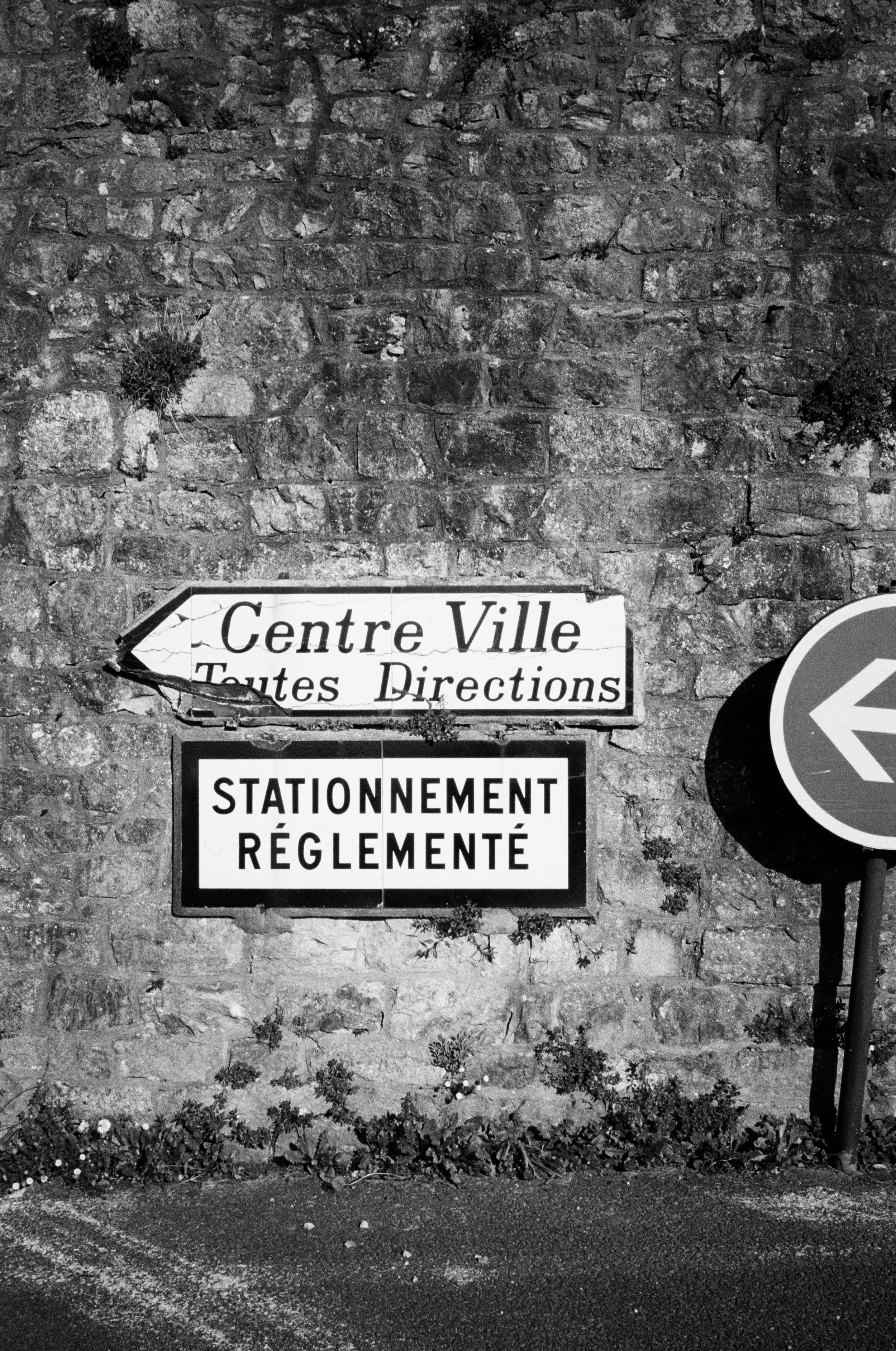 Black-and-white photo of a brick wall with two traffic signs attached. The top sign is worn and points left, reading 'Centre Ville Toutes Directions' in French. The bottom sign is in French and reads 'Stationnement Réglementé', indicating regulated p
