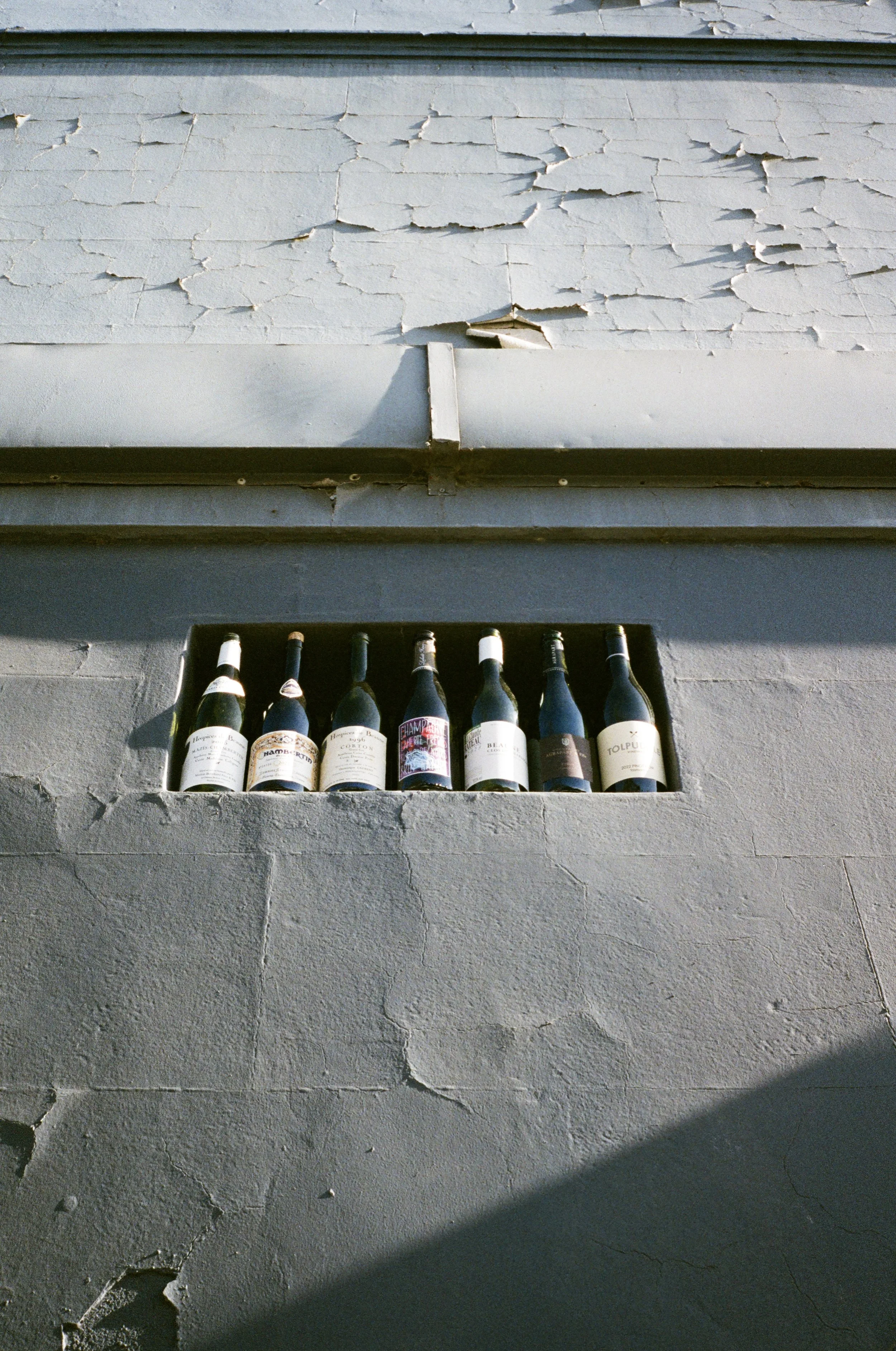 A row of wine bottles placed in a niche in a gray, cracked wall with peeling paint.