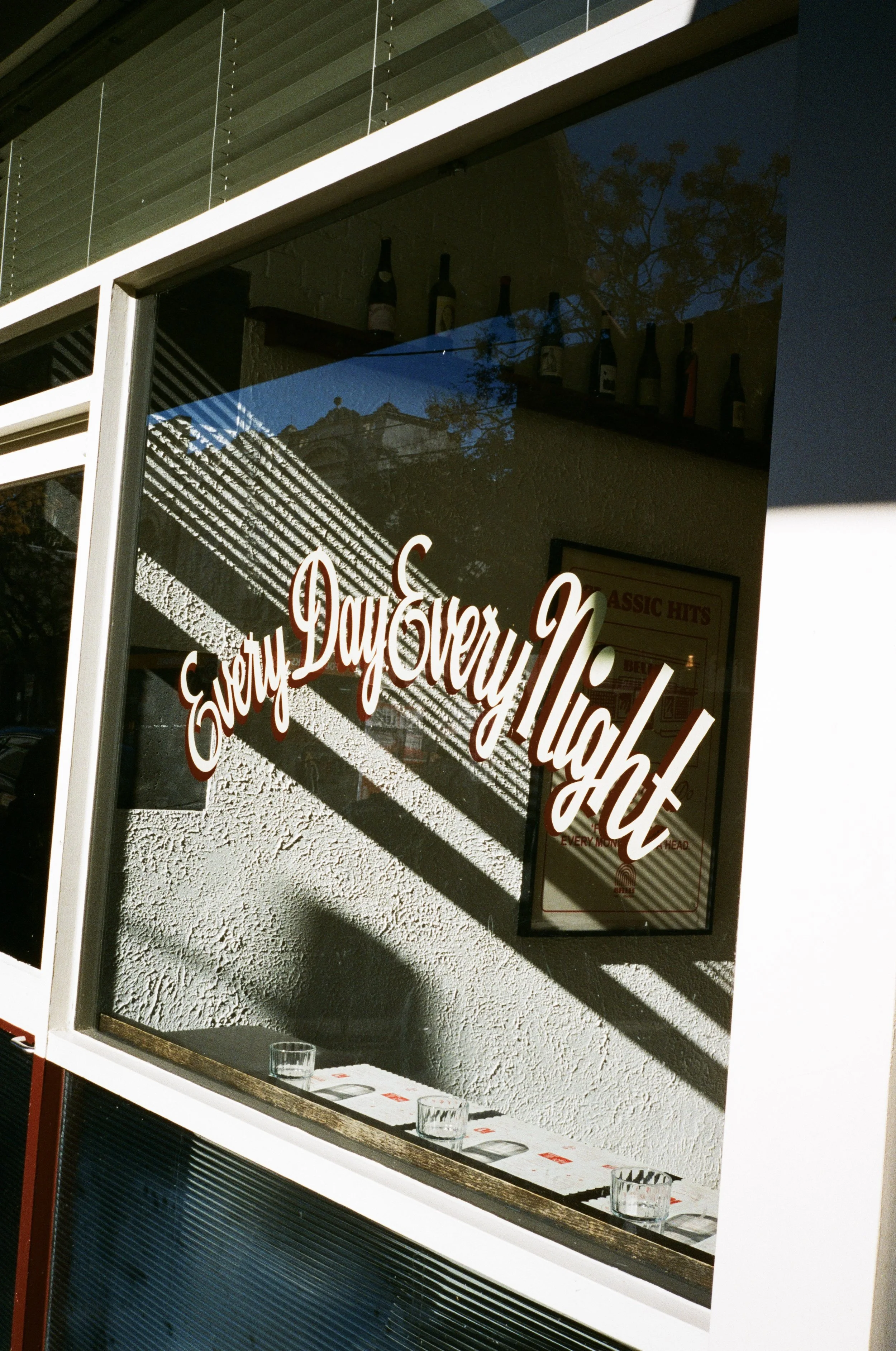 A storefront window with red cursive writing that says "Sorry Every Night" and displays bottles on a shelf inside. Reflections of a building and trees are visible on the glass.