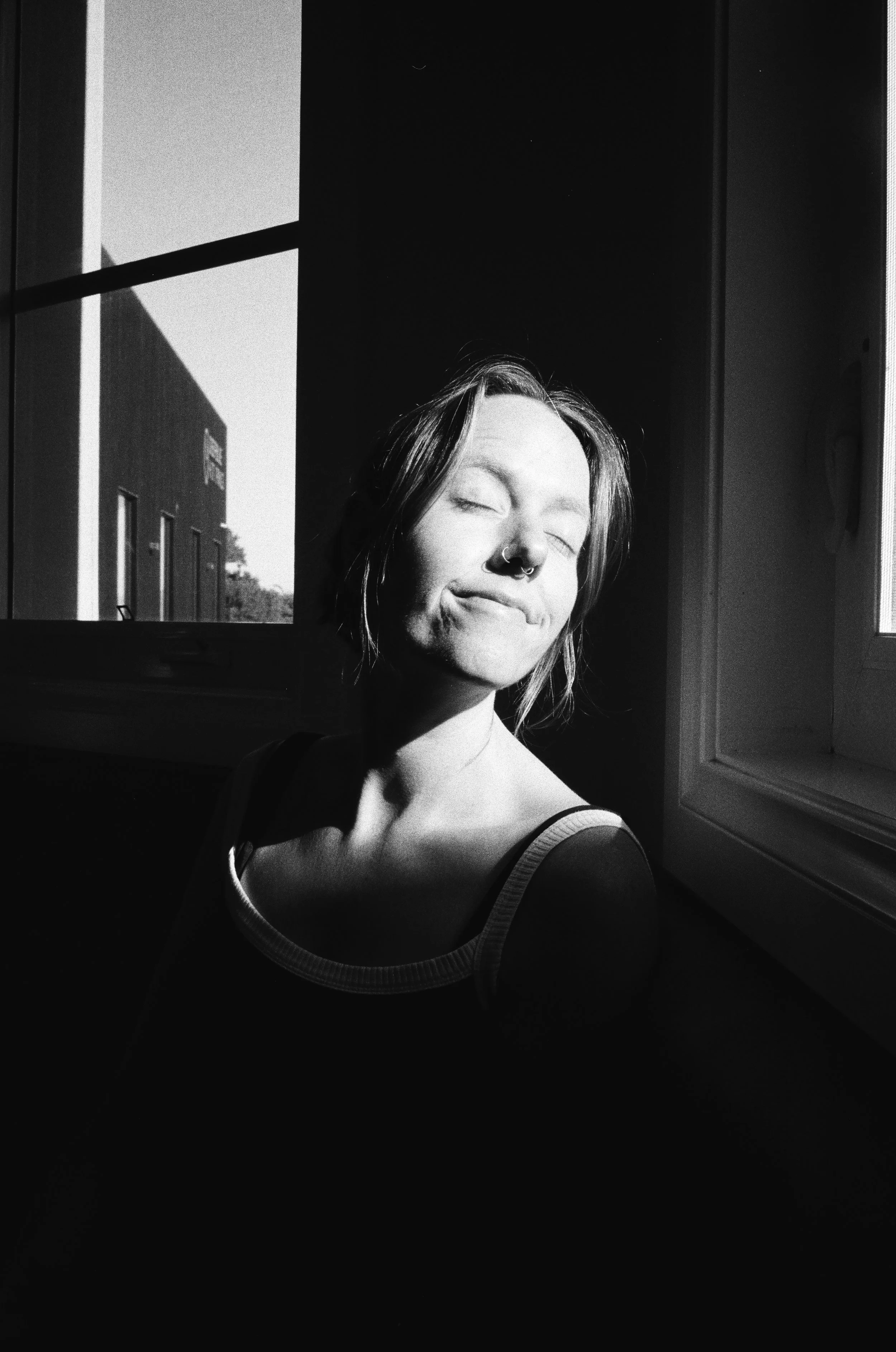 A young woman with closed eyes smiling in black and white, with sunlight illuminating her face from a window behind her.