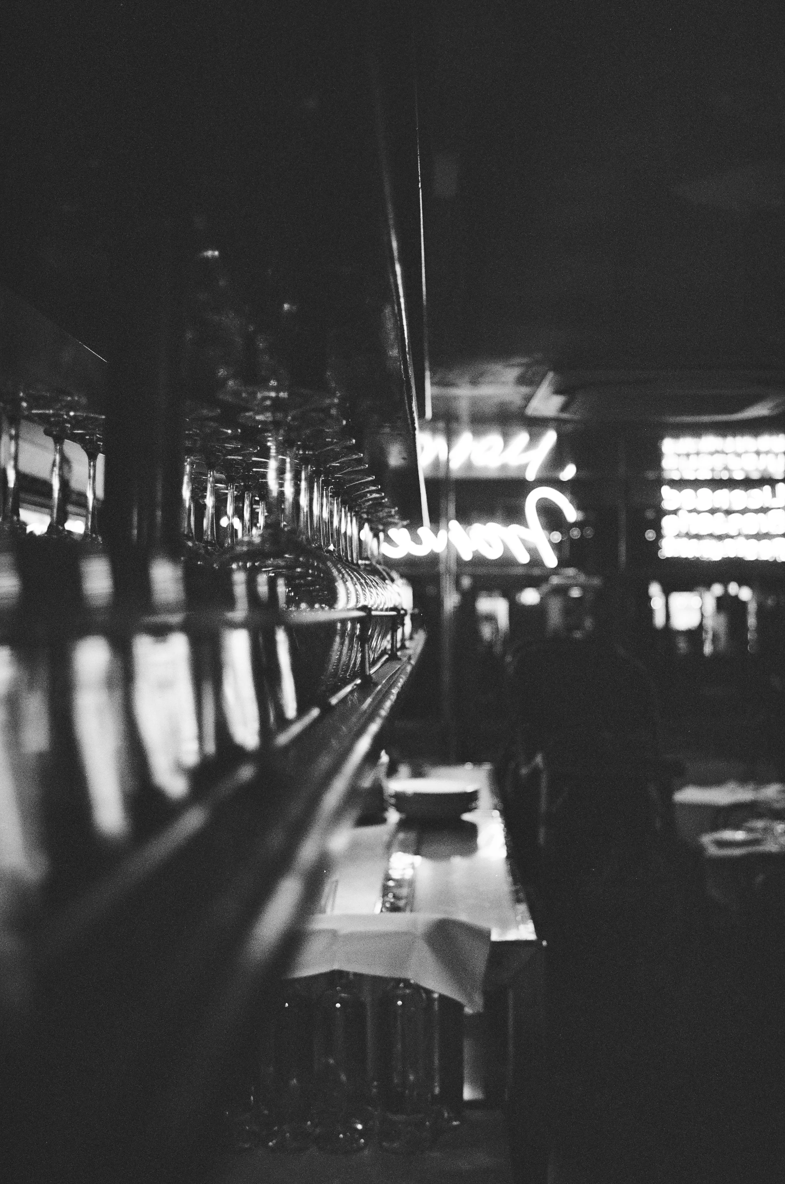 Black and white photo of a row of upside-down glasses on a bar shelf with neon signs in the background.