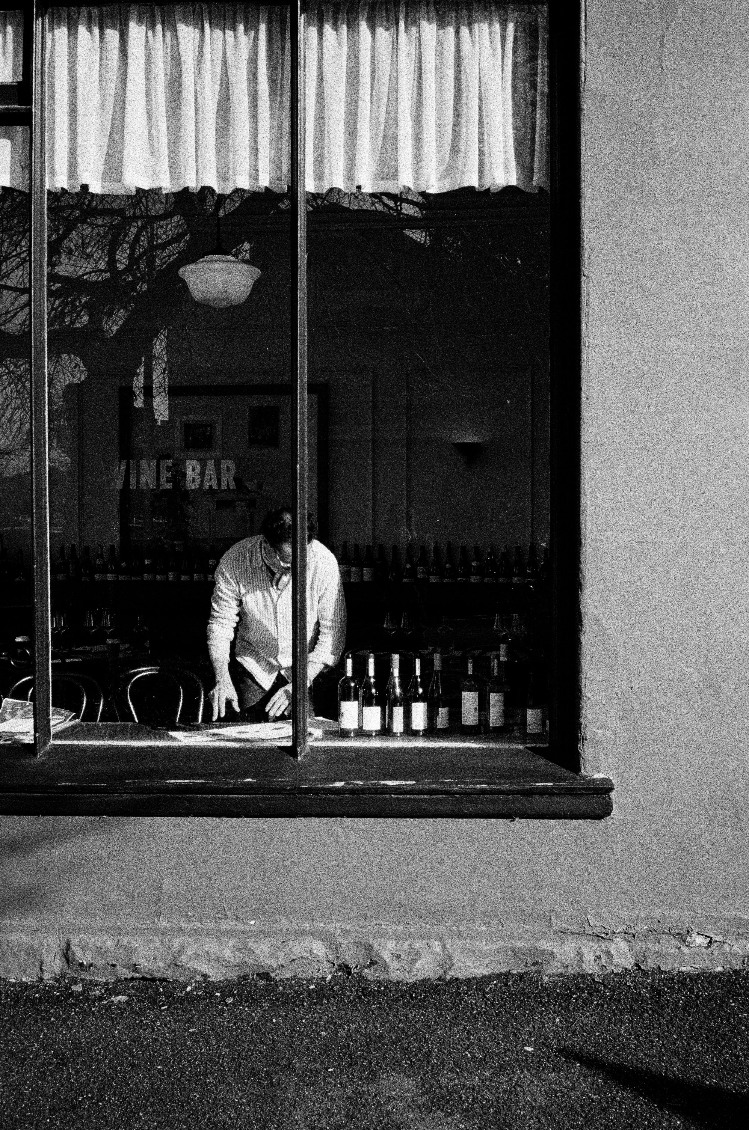 A man with a mask, shirt, and tie arranging bottles of wine on a counter inside a wine bar, seen through a window with curtains and reflections of trees and building exterior.