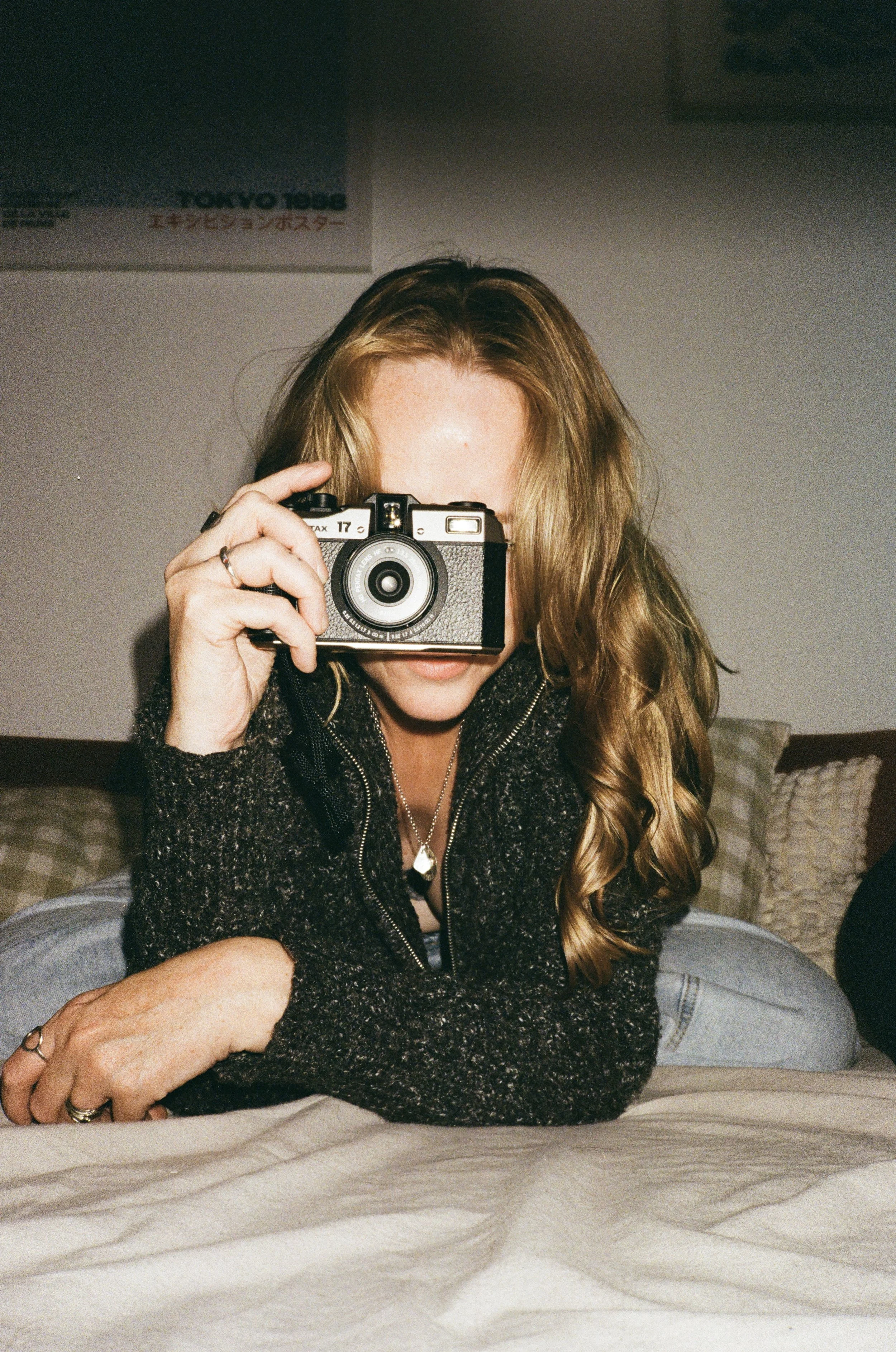 Person with long curly hair holding a camera in front of their face, sitting on a bed with a blanket, in a room with pillows and a poster on the wall.