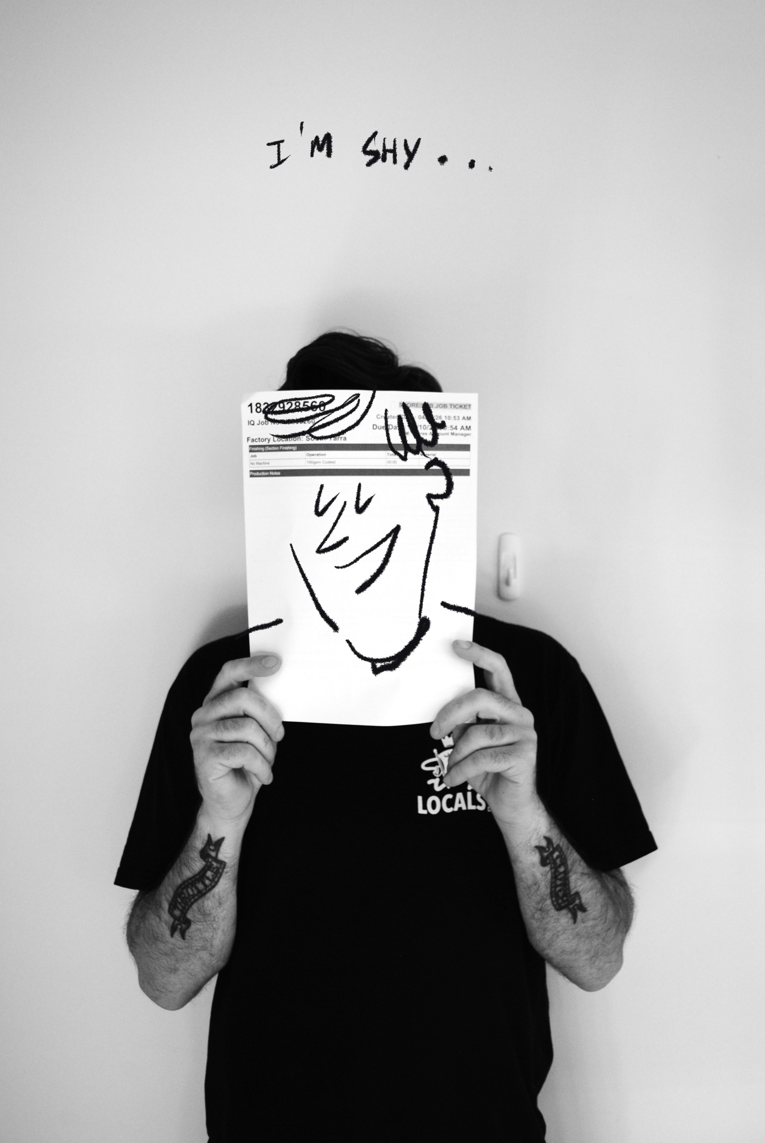 Person with tattoos on arms holding a paper with a drawn face in front of their face, standing against a white wall with the words "I'm SHY..." written above.