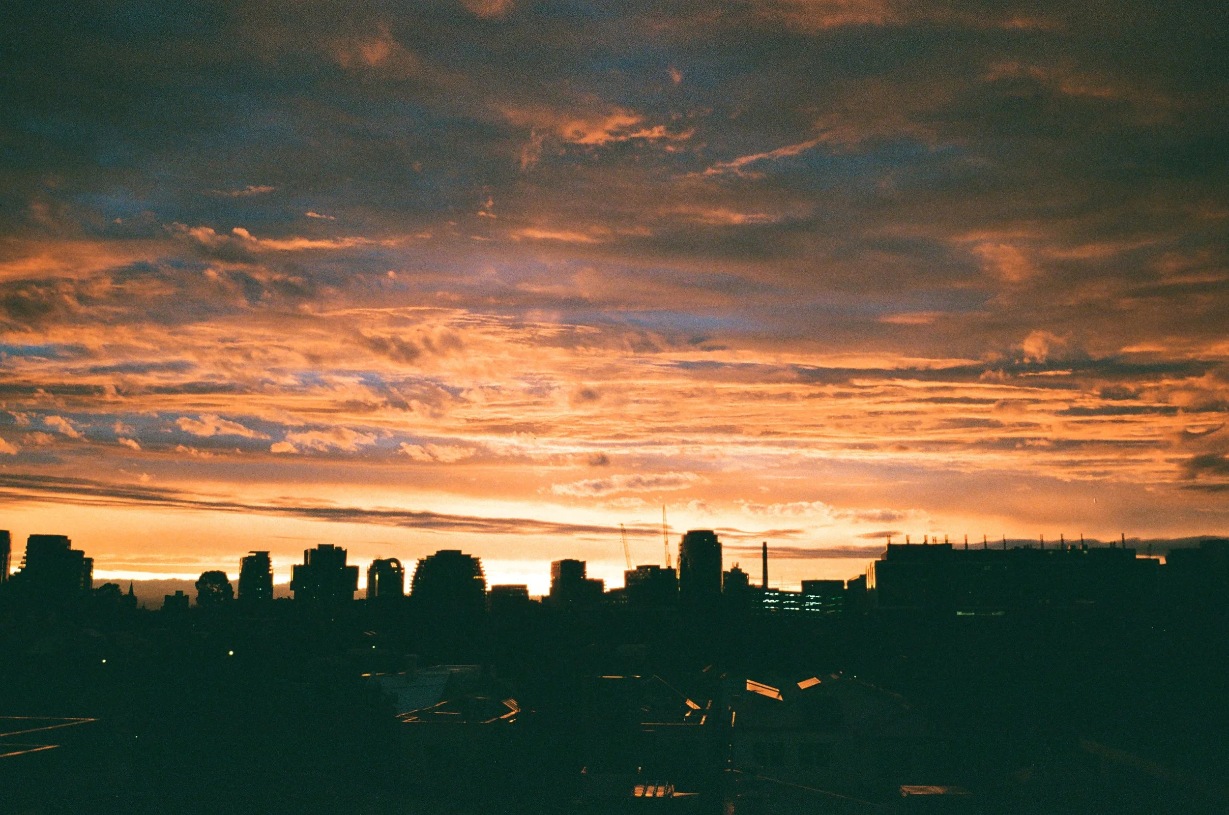 A city skyline at sunset with dark silhouette buildings and a colorful sky filled with clouds in shades of orange, pink, and purple.