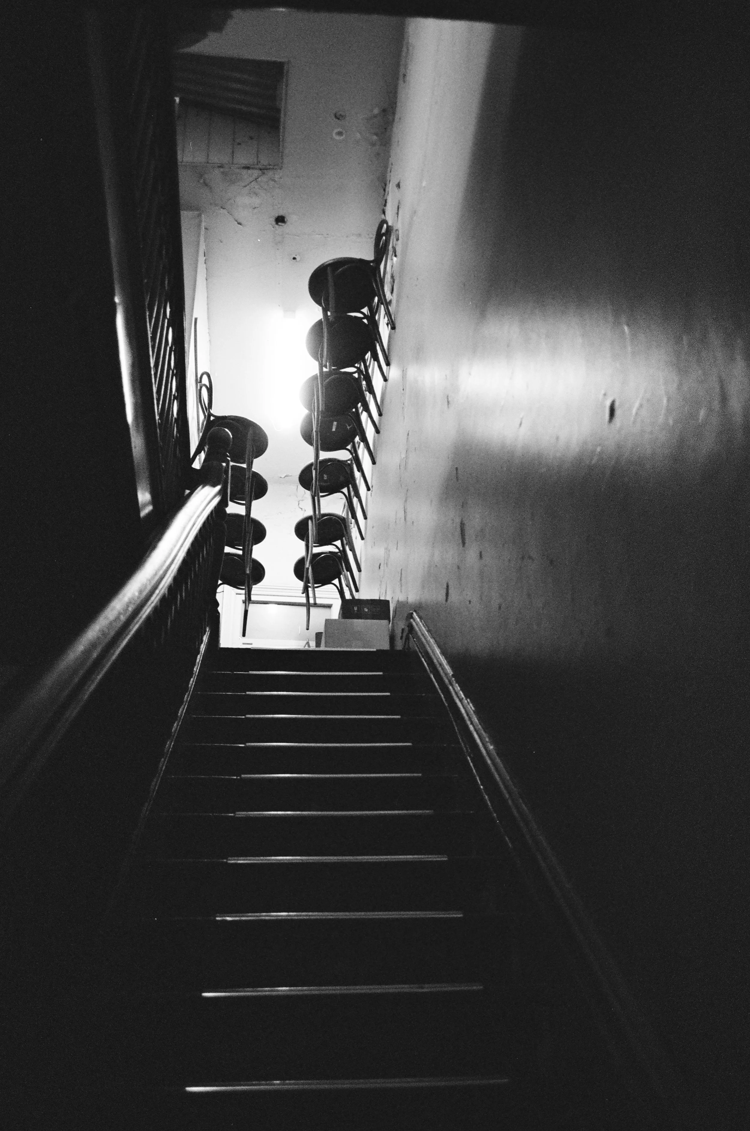 A staircase leading up to a room with a wooden floor and wall. On the side, there are multiple chairs stacked against the wall. The room appears to be in disrepair, with peeling paint and cracks visible on the ceiling and wall. There is a bright ligh