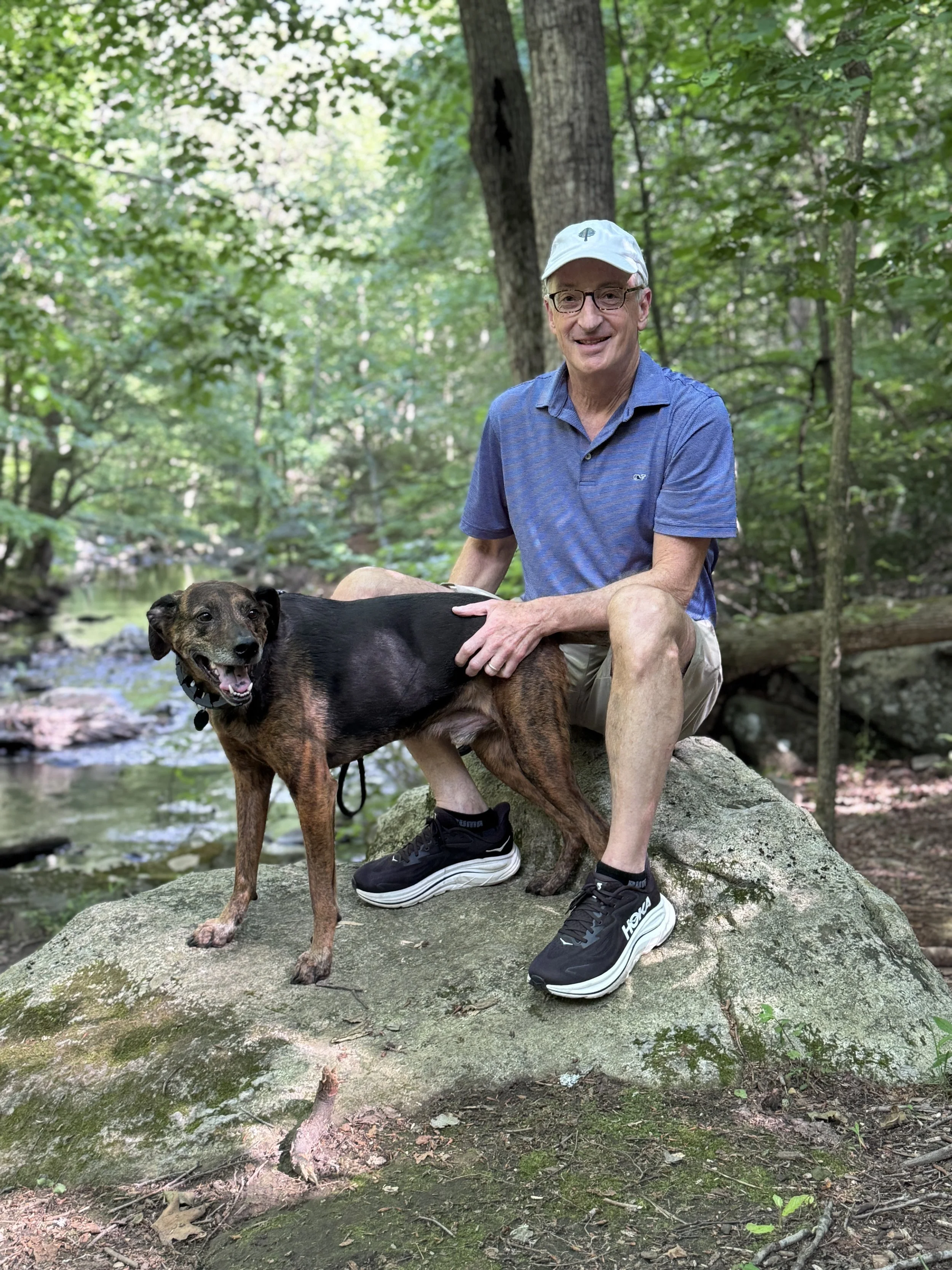 The author sitting on a large rock in a lush, green forest near a creek, accompanied by his happy hound loyally standing next to him.