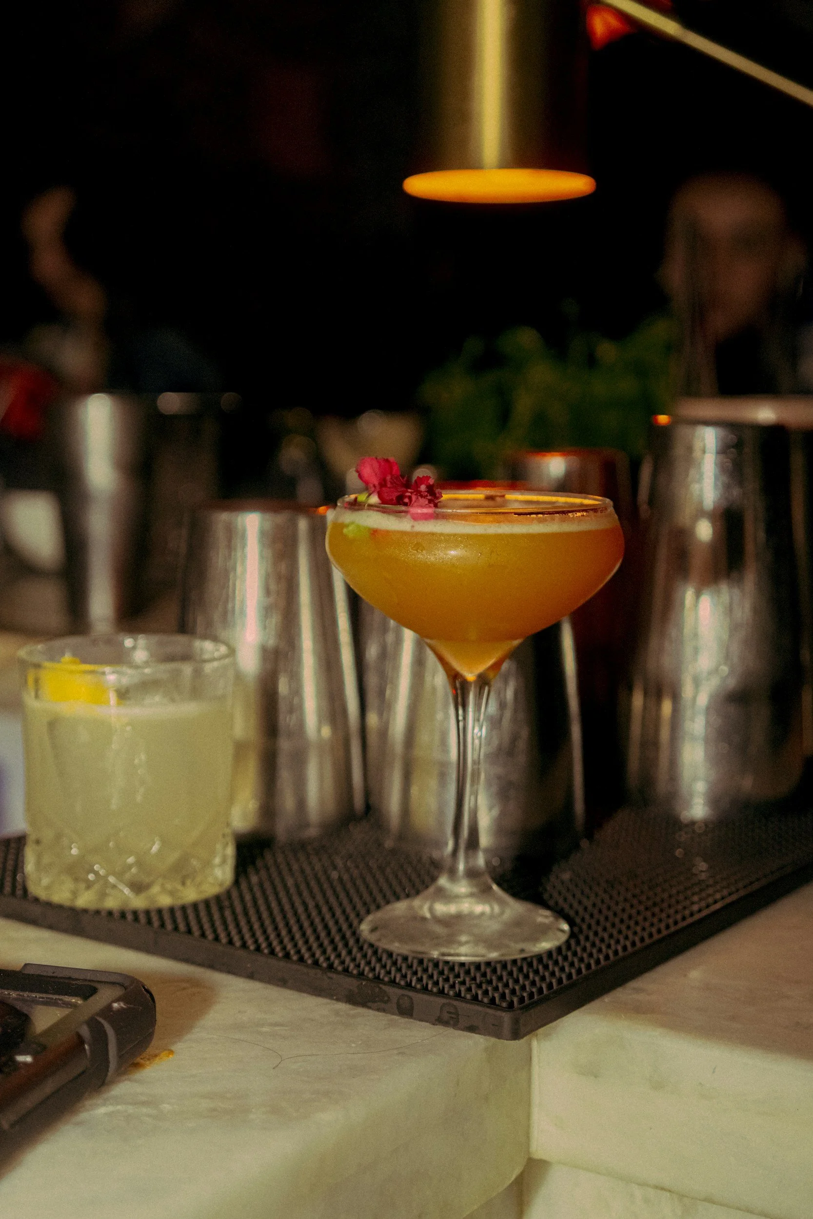 A yellow cocktail in a coupe glass garnished with a red flower sits on a bar counter. The background features blurred bar tools and equipment.