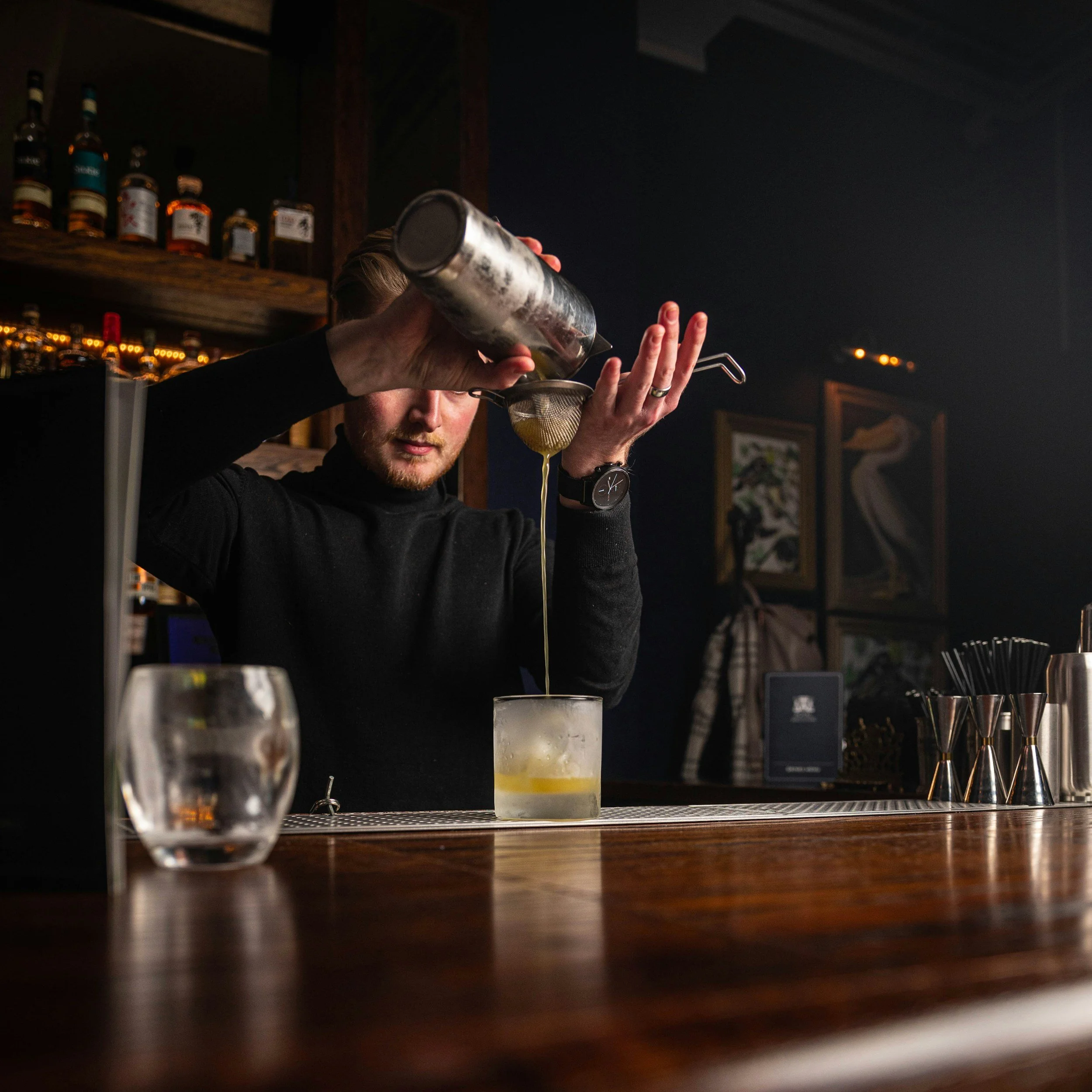 A bartender pouring a drink through a strainer into a glass at a bar, with liquor bottles on shelves in the background.