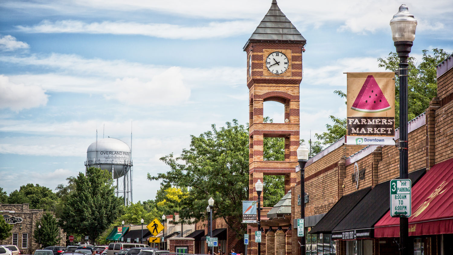 Downtown Main Street scene with a brick clock tower, shops, trees, cars, street signs, and a water tower in the background.