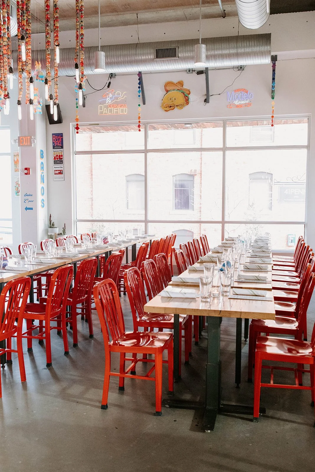 A bright restaurant dining room with red chairs and tables beside large windows.