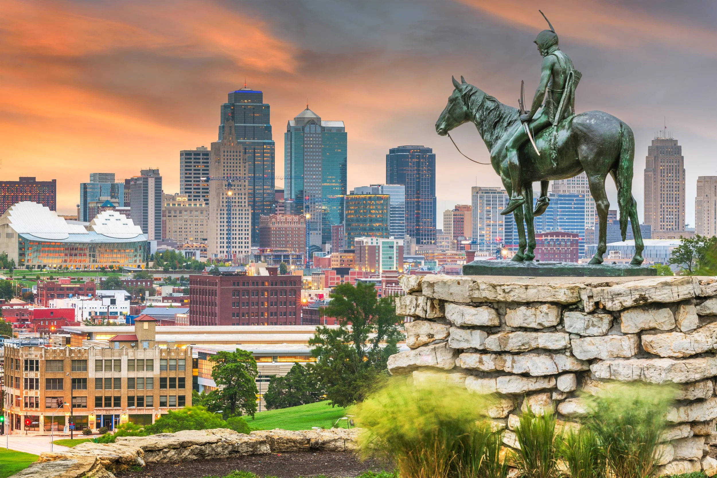 A bronze statue of an indigenous man on horseback, holding a bow and arrow, positioned on a stone platform overlooking a city skyline at sunset. The background features tall skyscrapers and a colorful sky with orange and pink hues.