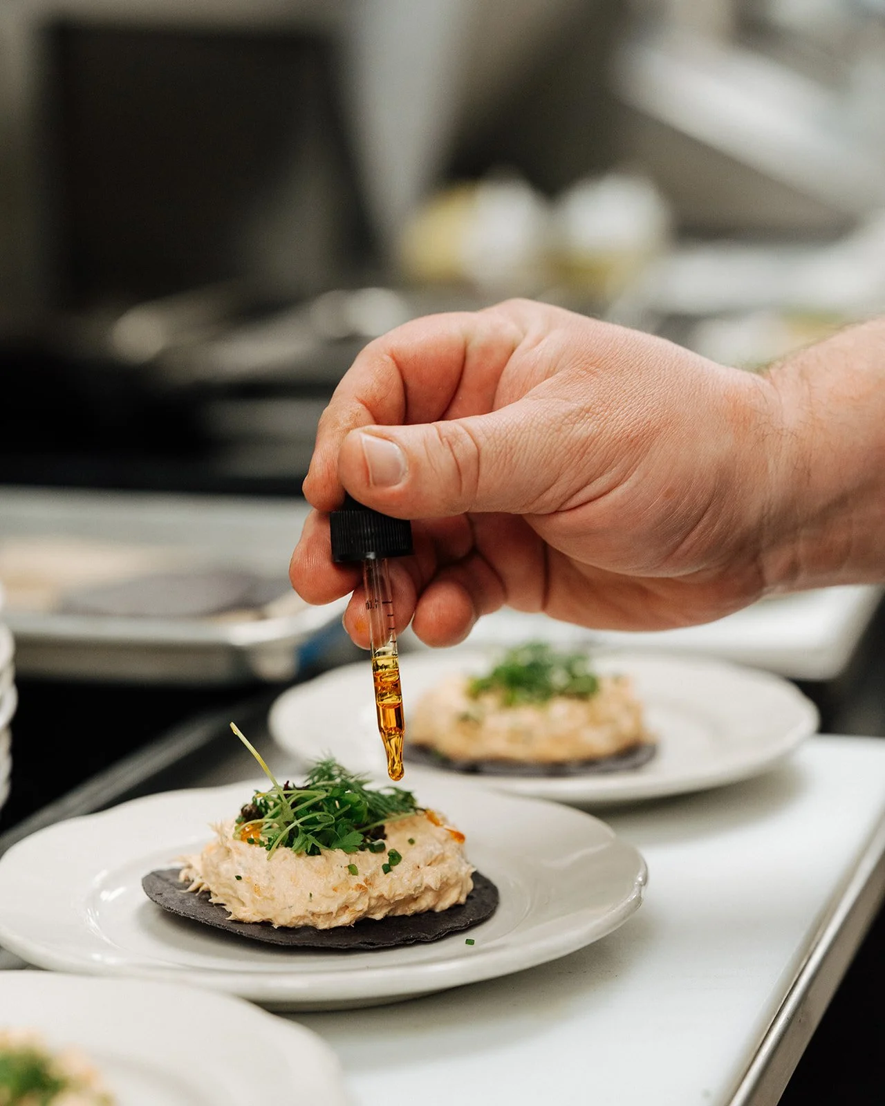Person using a dropper to add liquid onto a dish topped with herbs in a professional kitchen.
