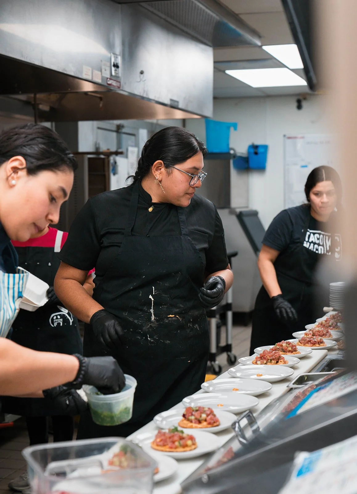 Women preparing and plating dishes in a commercial kitchen, with plates of food on a counter.