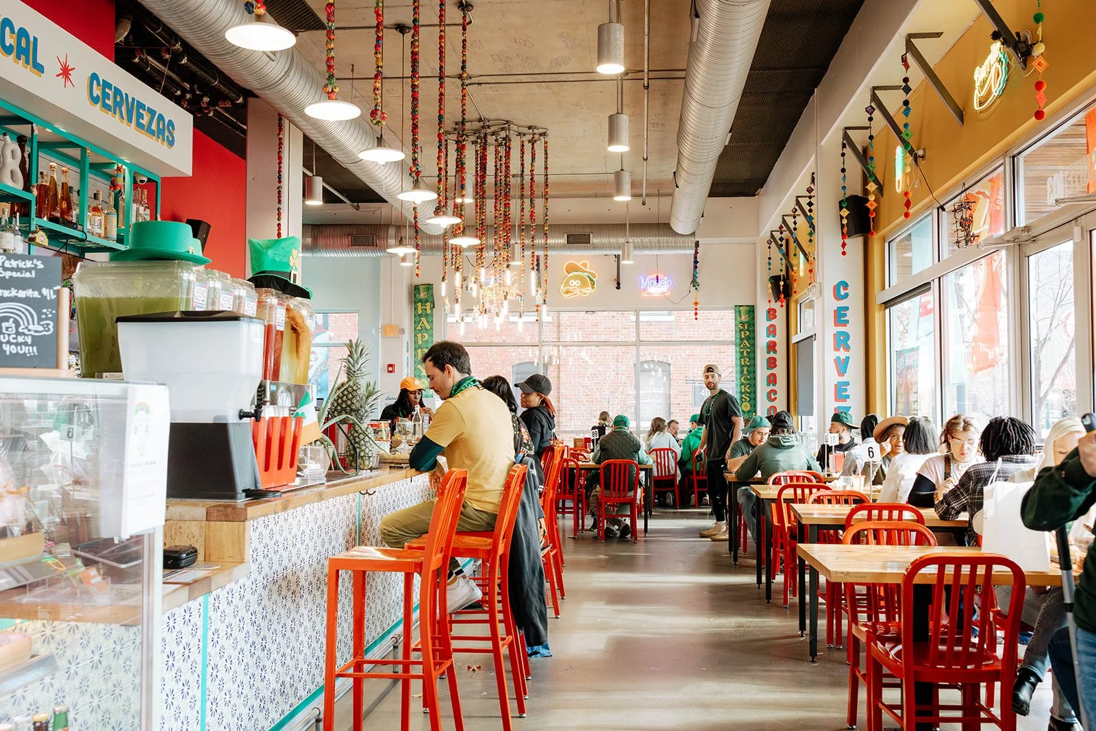 Inside a lively cafe with bright red chairs, patrons sitting at tables, and a counter where a man is seated. The decor features colorful hanging garlands, neon signs, and large windows letting in natural light.