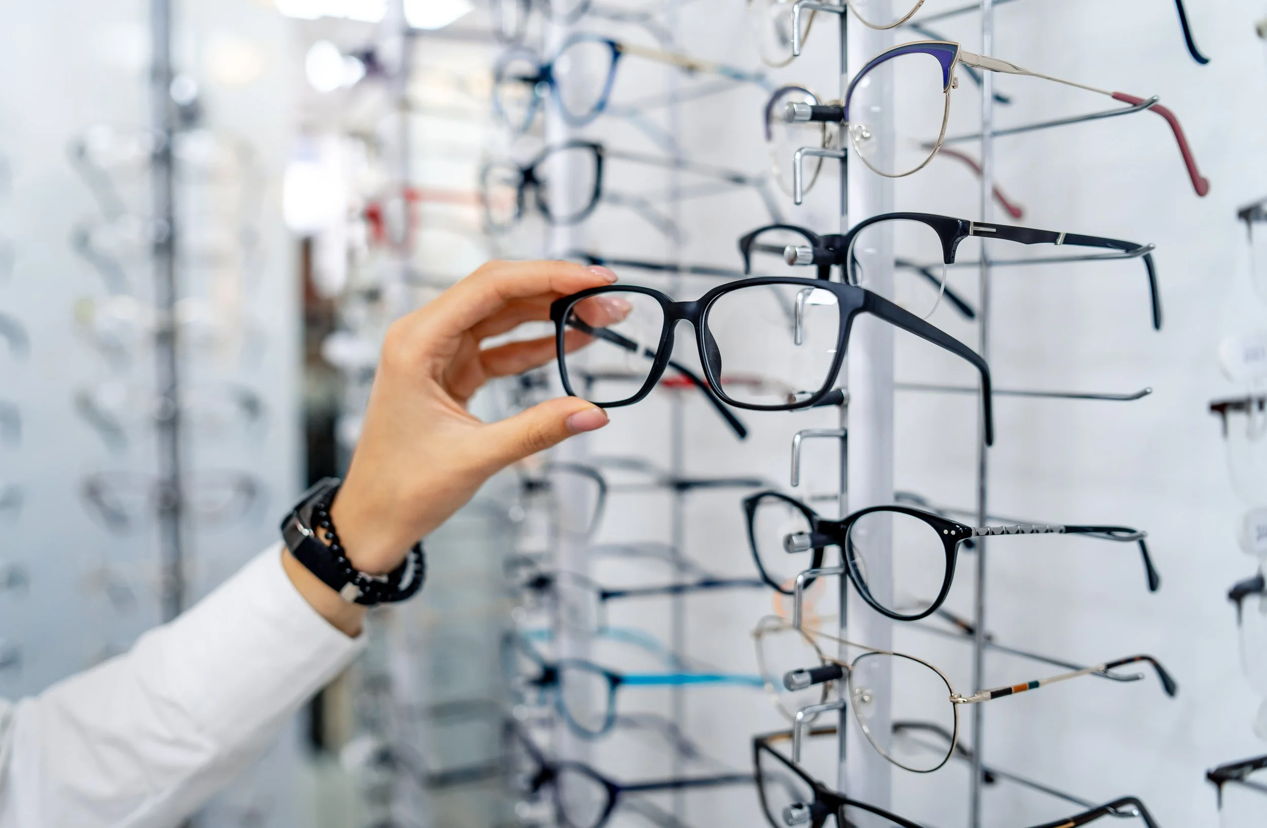 A person wearing a white long-sleeve shirt and a black bracelet on their left wrist is holding a black eyeglass frame in front of a display of various eyeglasses on a wall.