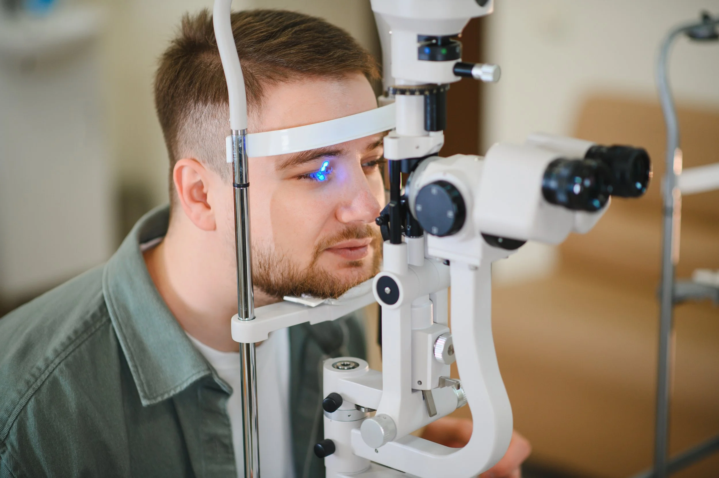 A young man with brown hair and a beard is using a slit lamp at an eye examination. He is looking into the device, which is used for eye health assessments, while the doctor or technician adjusts the equipment.