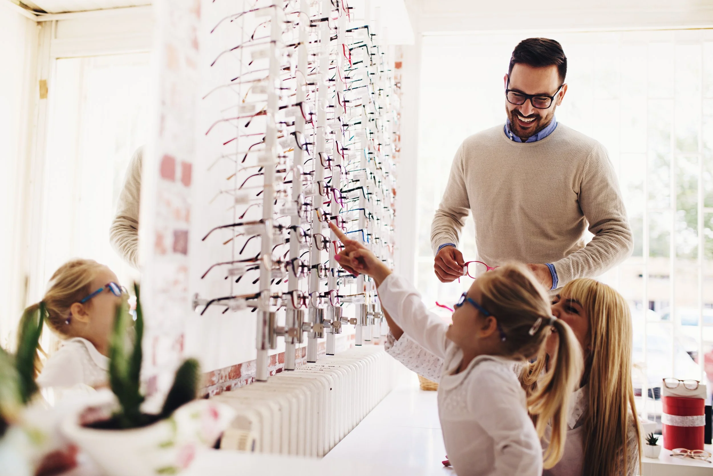A man with glasses and a beige sweater smiling at children trying on glasses inside an optical store.