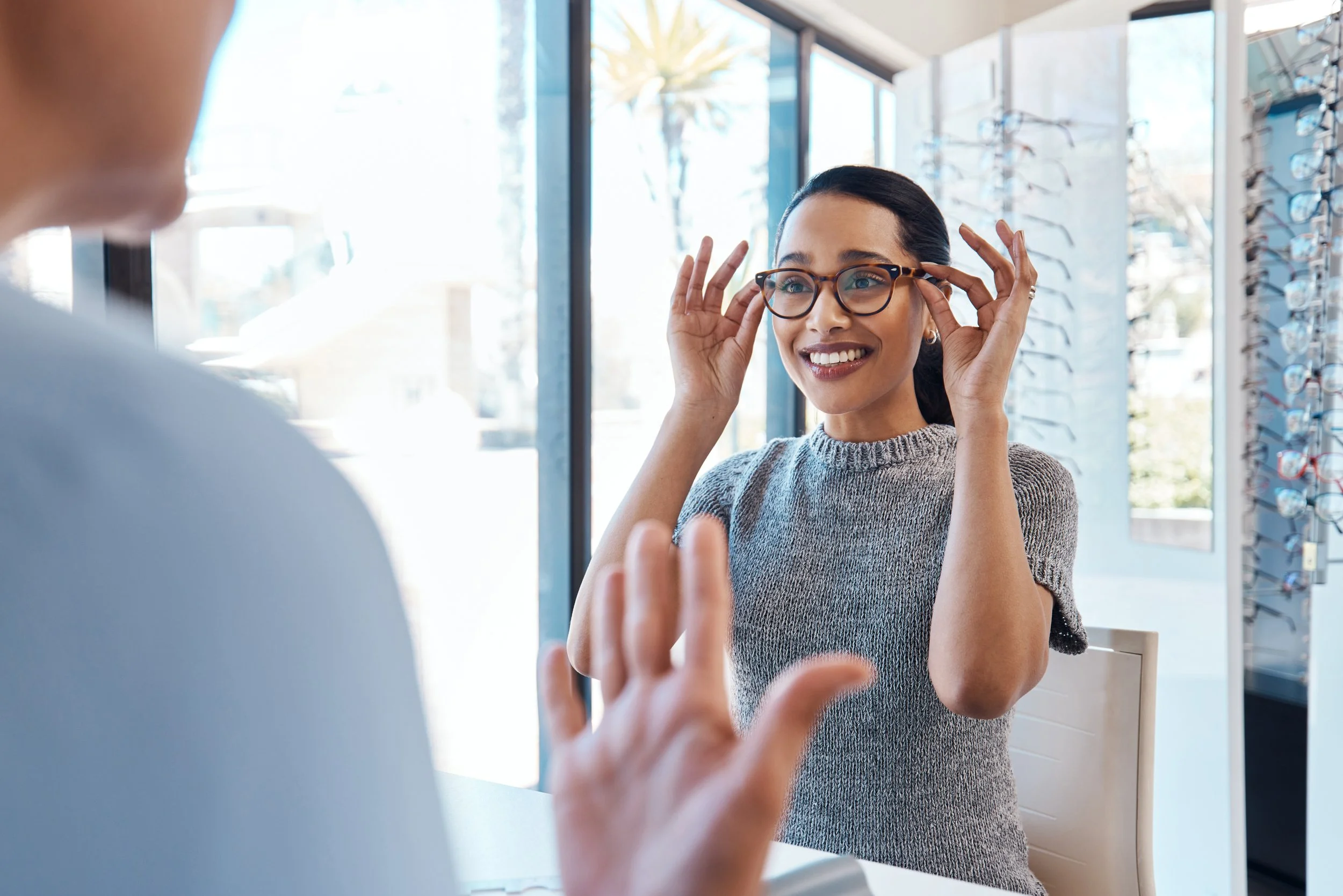 A woman with dark hair and glasses trying on a pair of eyeglasses at an optical store, smiling and holding the glasses with both hands.