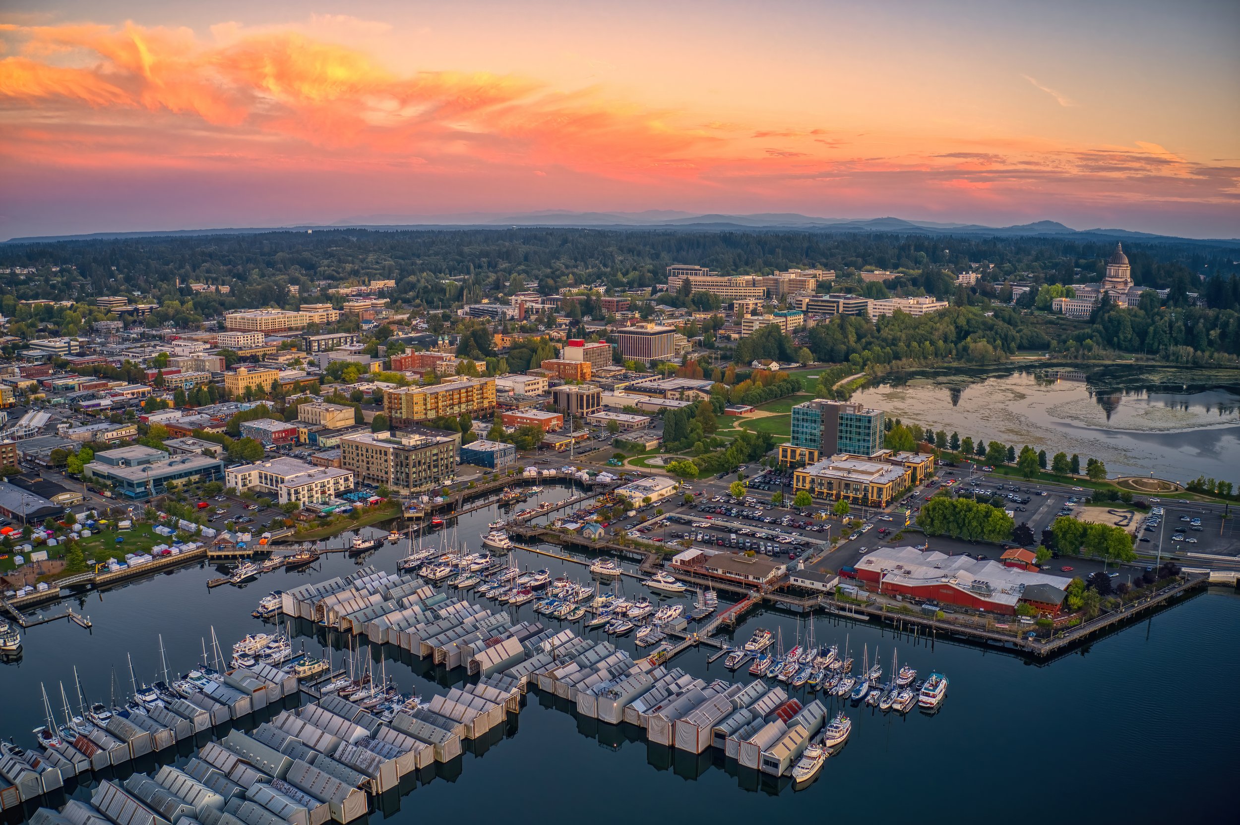 Aerial view of a city skyline at sunset with a body of water and marina in the foreground, and a mix of modern buildings and greenery in the background, including a domed government building.