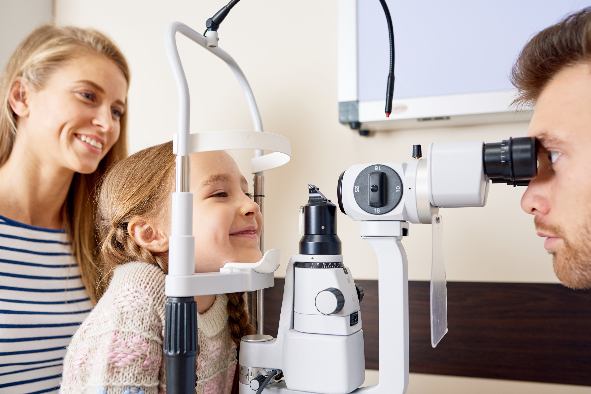 An eye doctor examining a young girl with a slit lamp, while a woman, possibly her mother, smiles nearby.