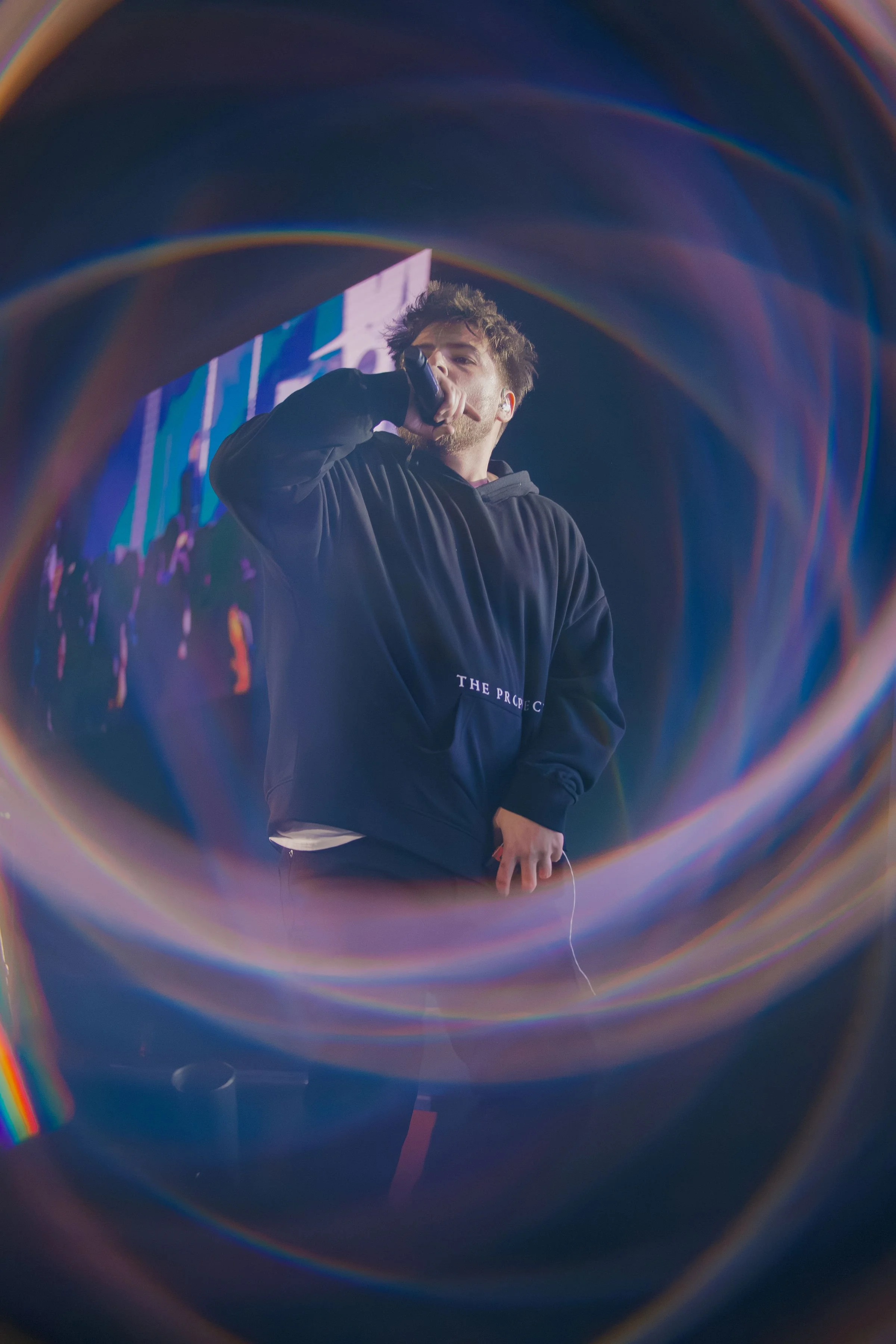 A young man with brown hair and a beard is holding a microphone and performing on stage, visible through a colorful, circular light pattern.
