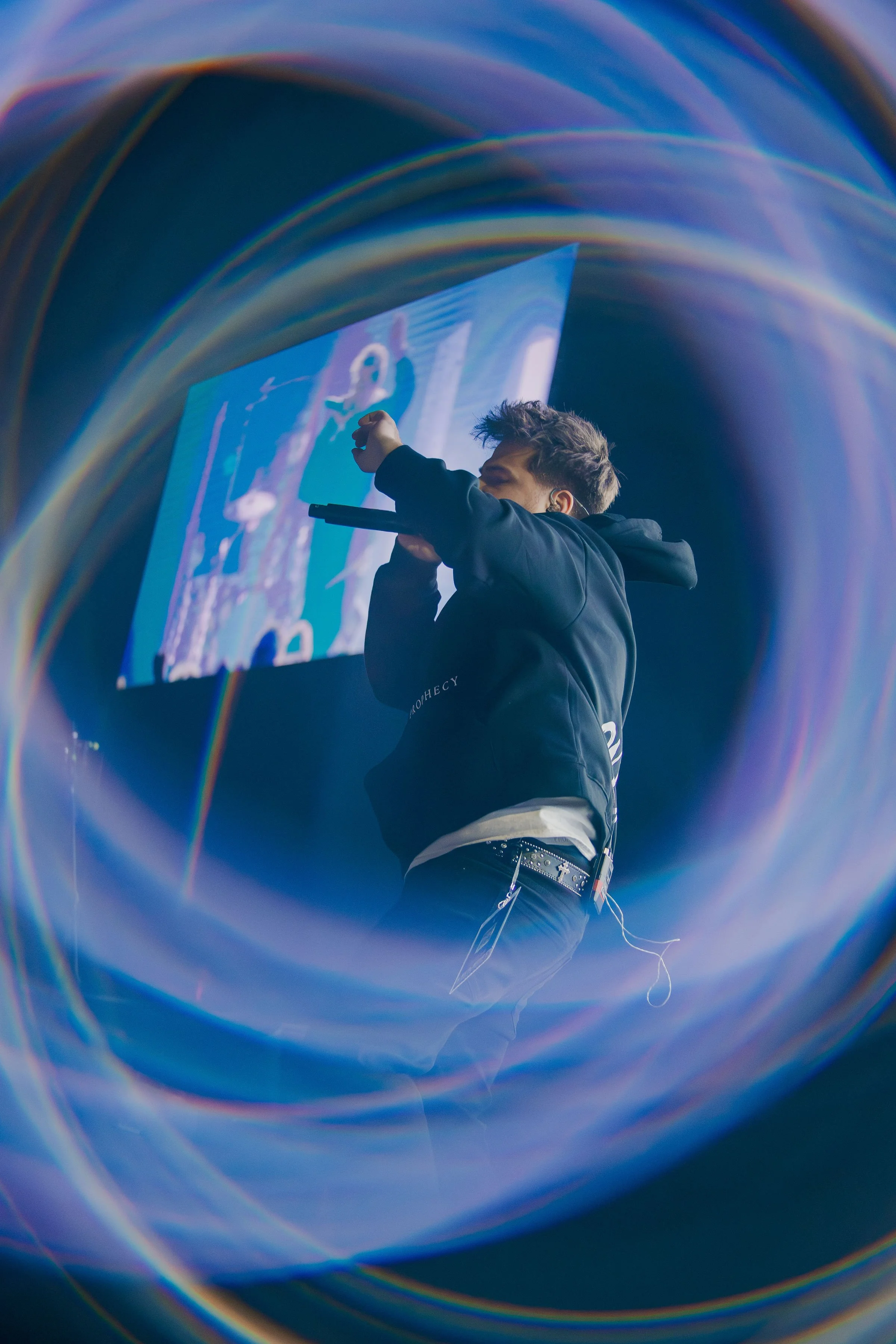 A person with short, dark hair is performing on stage, holding a microphone close to their mouth, with a large screen behind them displaying an animated or digital image. The scene is framed with a swirling, rainbow-colored light pattern or lens flare.