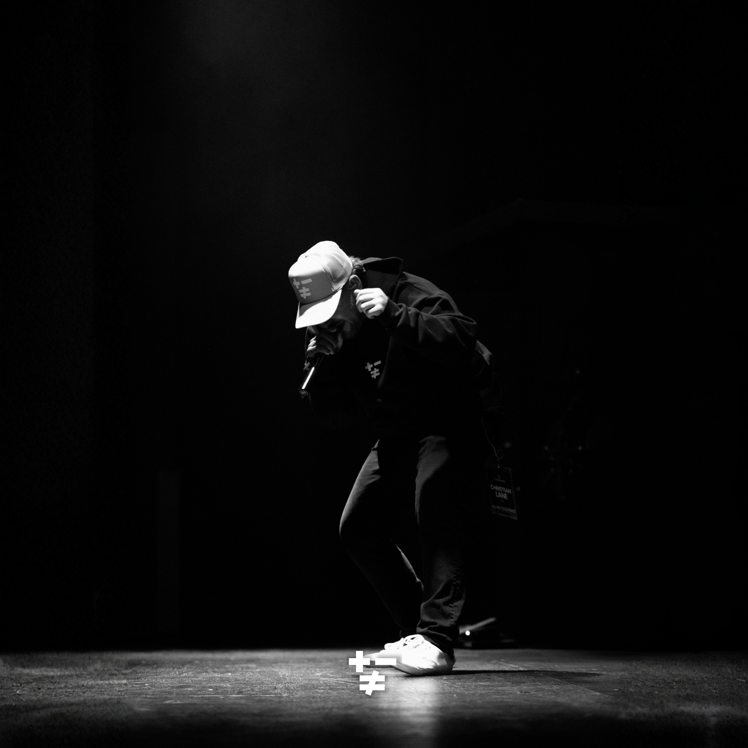 A person wearing a baseball cap and hoodie, crouching on a dark stage, with a large empty black background.