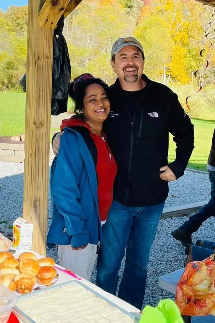 A woman and a man are smiling and posing together at an outdoor gathering, standing beside a table with baked goods and food. The background shows trees with autumn leaves.
