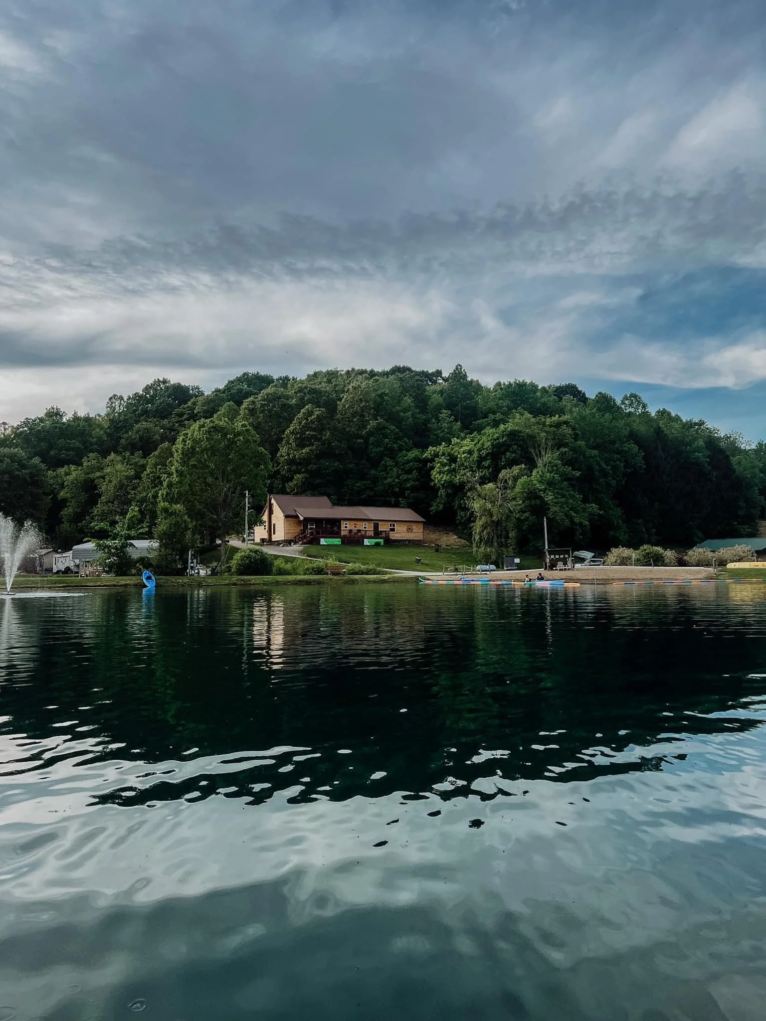 A lake with dark green water, surrounded by a lush forest on a hill, with a house at the base of the hill, a fountain, and colorful paddle boats on the shore. The sky is cloudy with patches of blue.