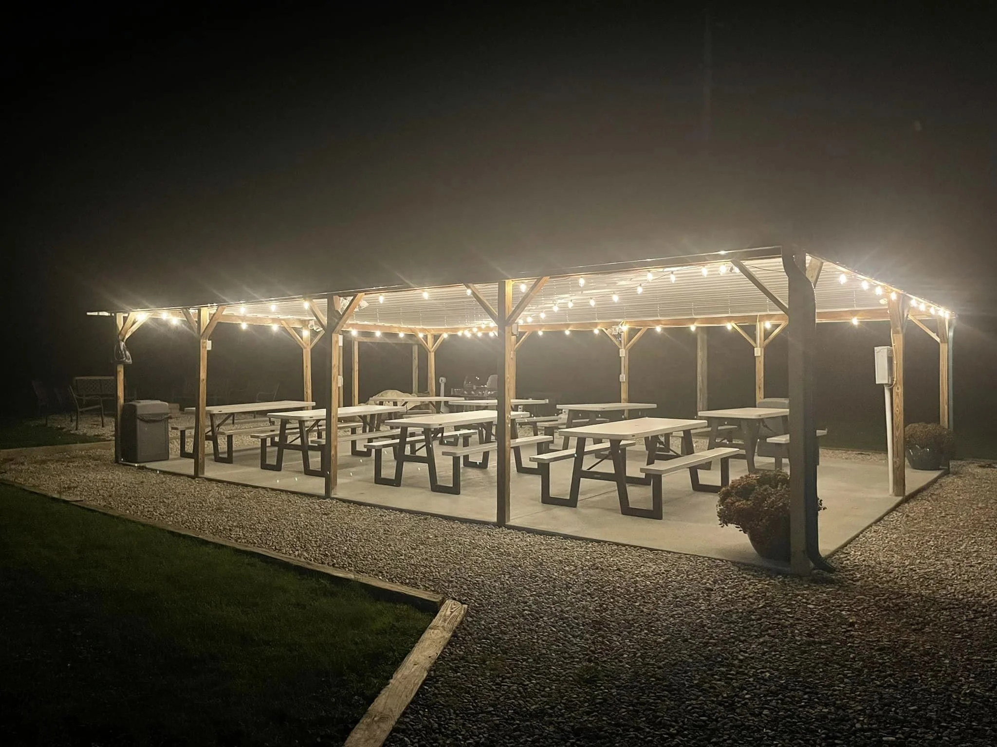 An outdoor pavilion at night with string lights hanging from a wooden structure, covering multiple picnic tables and benches on a concrete slab, surrounded by gravel and grass.