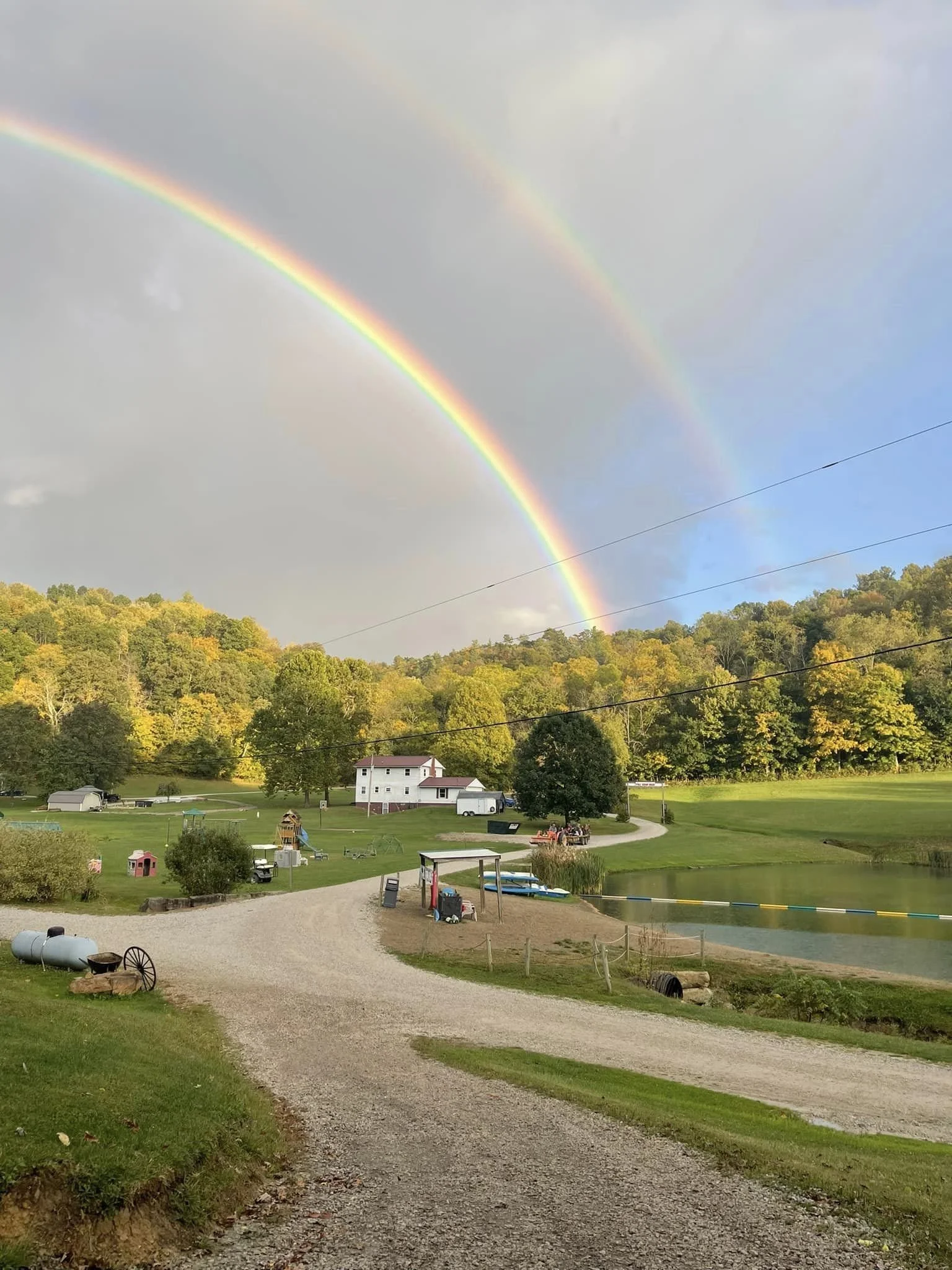 A scenic landscape with a gravel pathway leading to a grassy area containing trees, small structures, a pond, and a cart by the water, under a sky with double rainbow and partly cloudy weather.