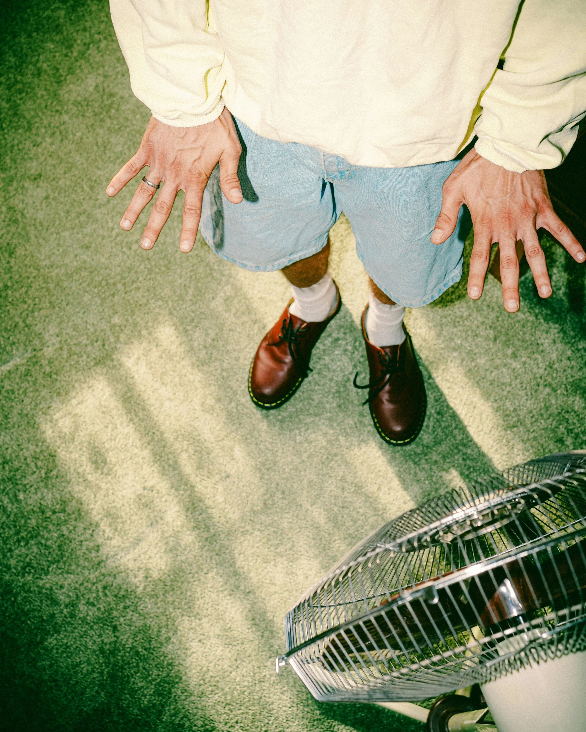A person wearing a white hoodie, light blue shorts, white socks, and brown boots standing on a green carpeted floor next to a metal fan.