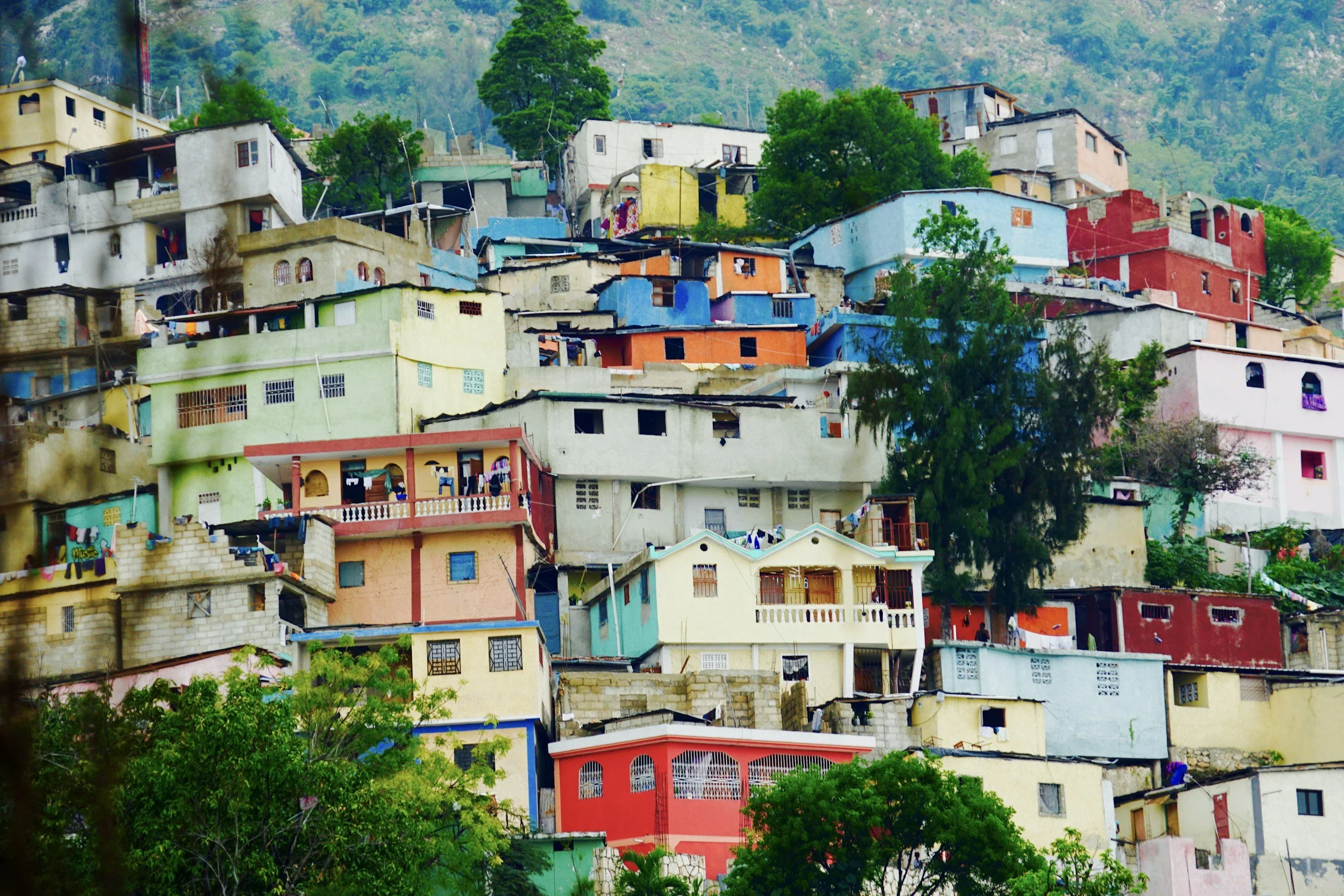 Colorful houses stacked on a hillside with green trees and a mountainous background.