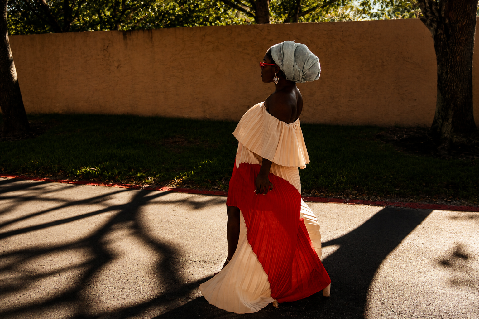 A woman walking outdoors on a sunny day, wearing a colorful pleated off-the-shoulder dress, a headwrap, and sunglasses, with trees and a beige wall in the background.