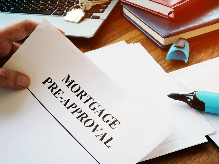 A person holding a paper titled 'Mortgage Pre-Approval' on a wooden desk with books, a pen, a highlighter, a blue mouse, and a laptop.