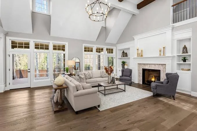 Bright living room with white walls, high vaulted ceiling, large windows, beige sectional sofa, dark armchairs, fireplace, wooden floors, and modern chandelier.
