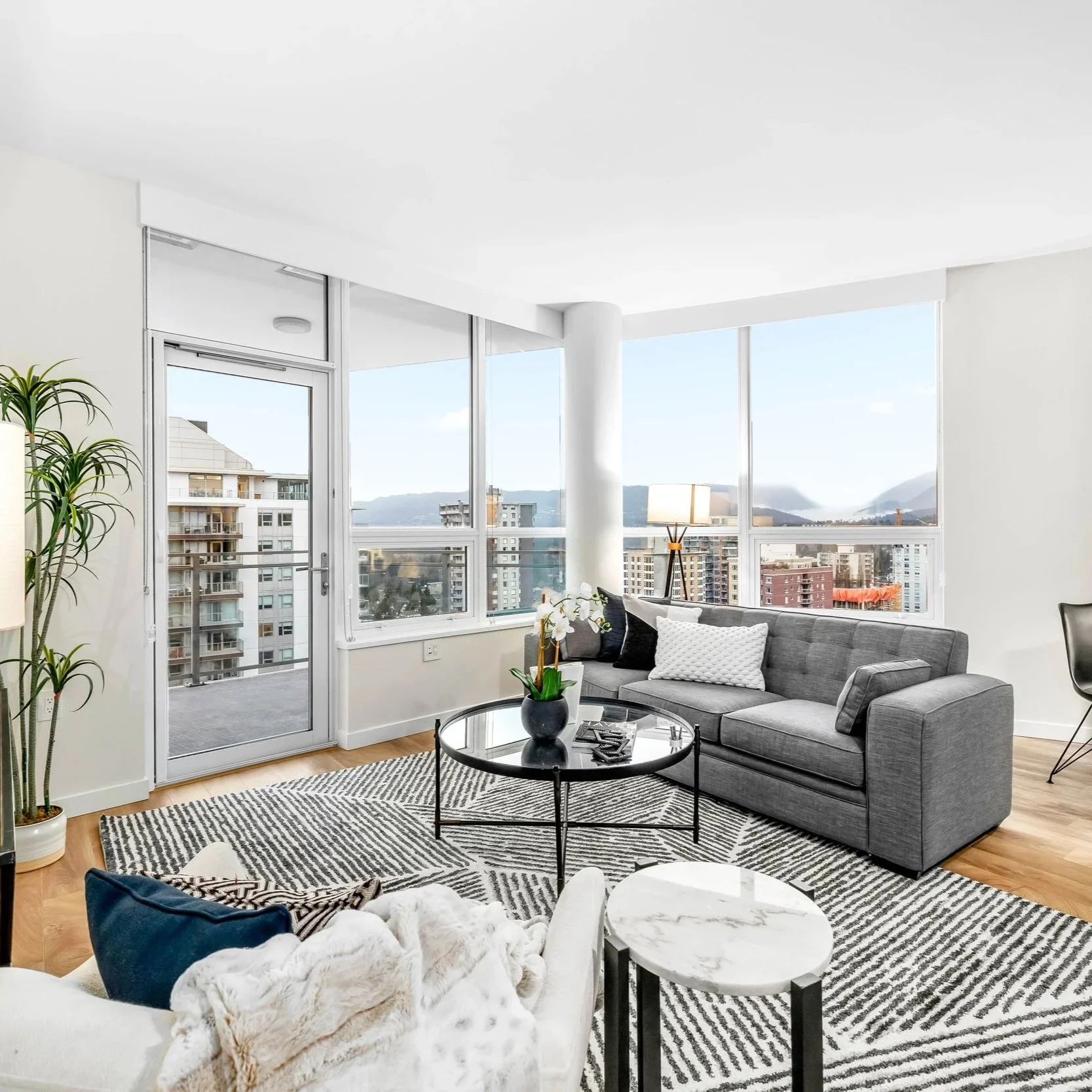 Modern living room with large windows and city view, gray sofa with pillows, black coffee table with a potted orchid, patterned rug, and a small round side table with a white marble top.