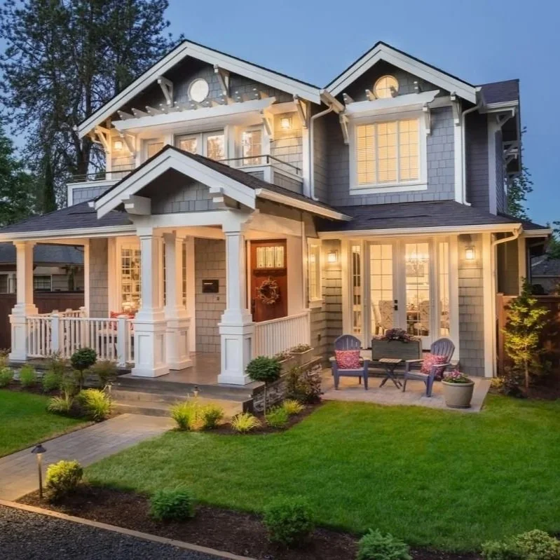 A two-story house with a porch, large windows, and a well-manicured lawn, illuminated with exterior lights at dusk.