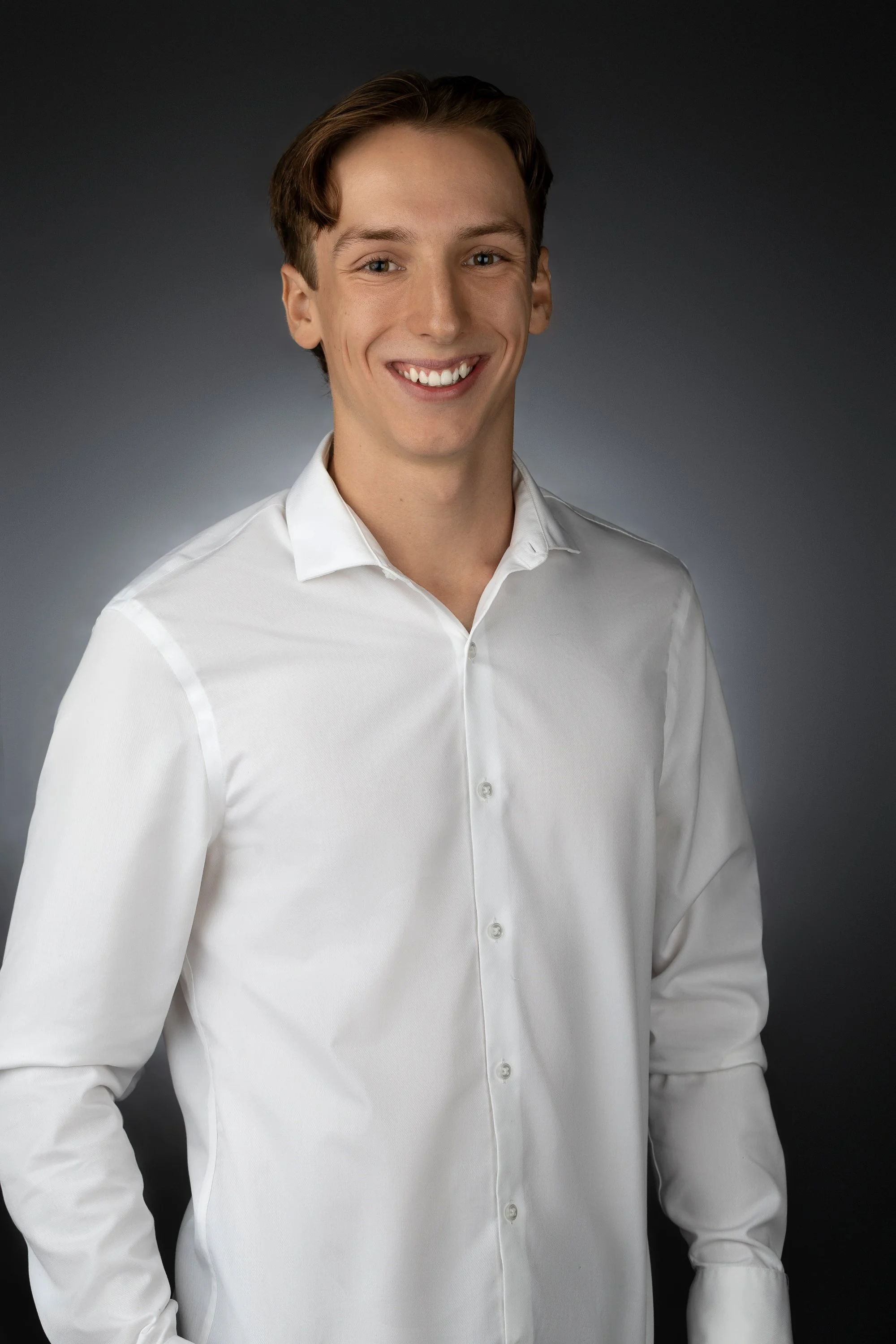 A young man with light skin, brown hair, and blue eyes, smiling, wearing a white dress shirt against a dark gradient background.