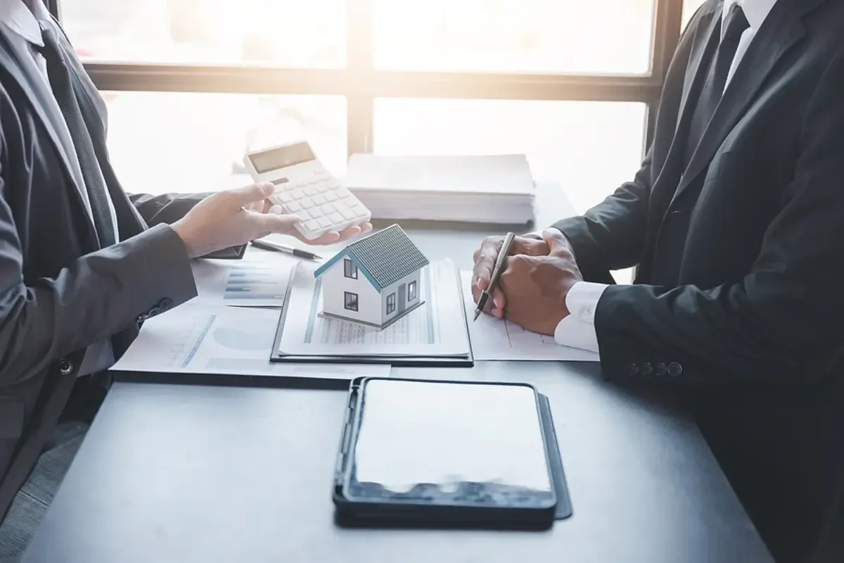 Two people in business suits sitting at a desk with a small house model, legal documents, calculator, and electronic tablet.