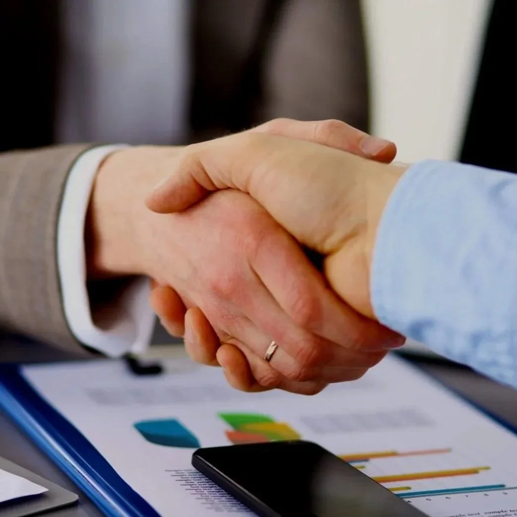 Close-up of two people shaking hands over a desk with a tablet, phone, and documents with graphs and charts.