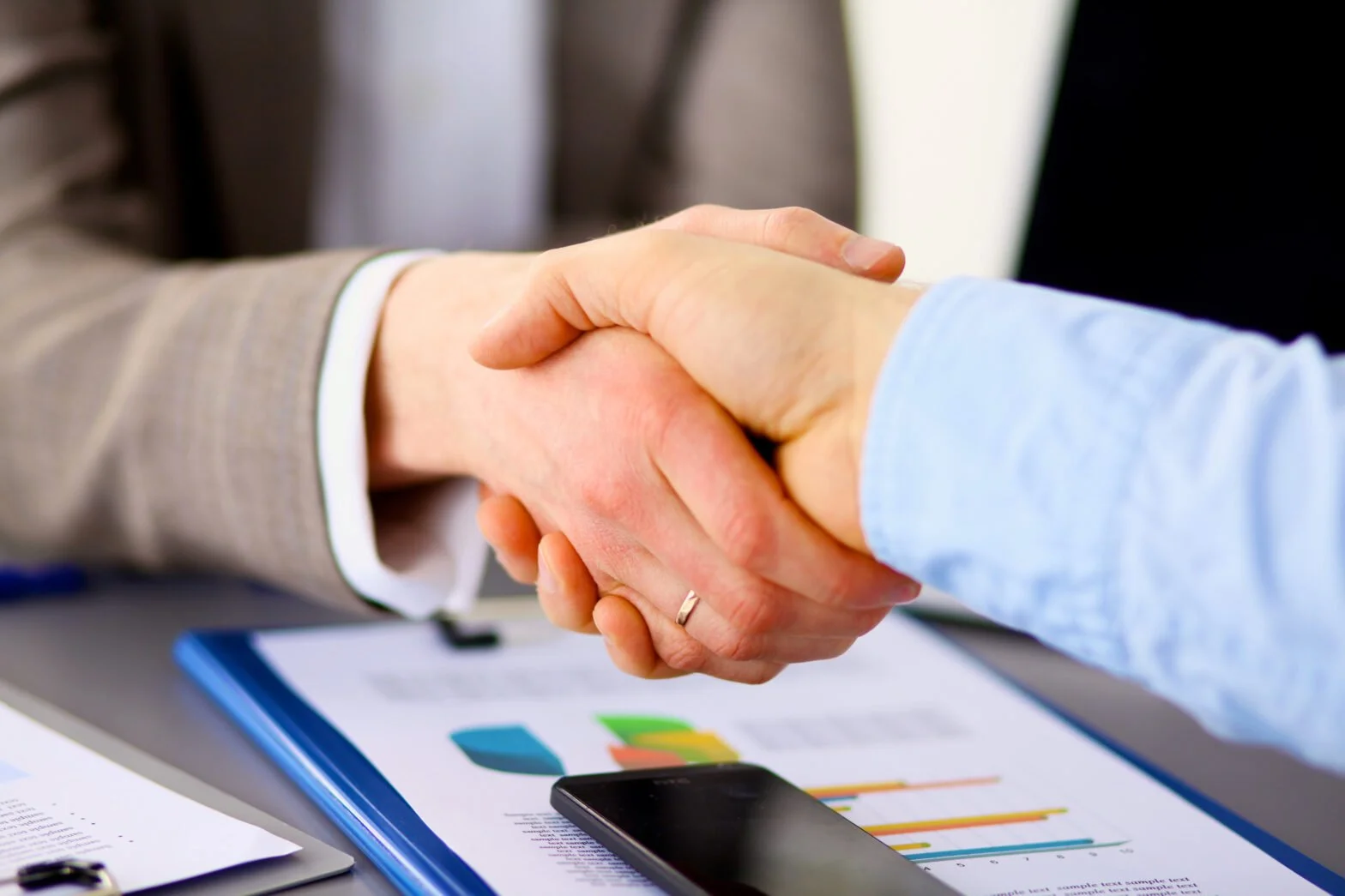 Close-up of two people shaking hands over a desk with charts, graphs, a smartphone, and papers.