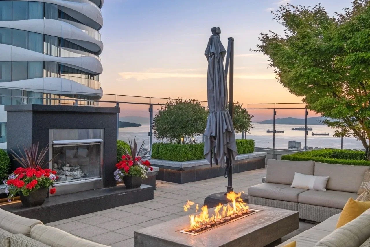A rooftop patio with outdoor seating, a small fire feature, potted flowers, and a view of the water and ships at sunset.