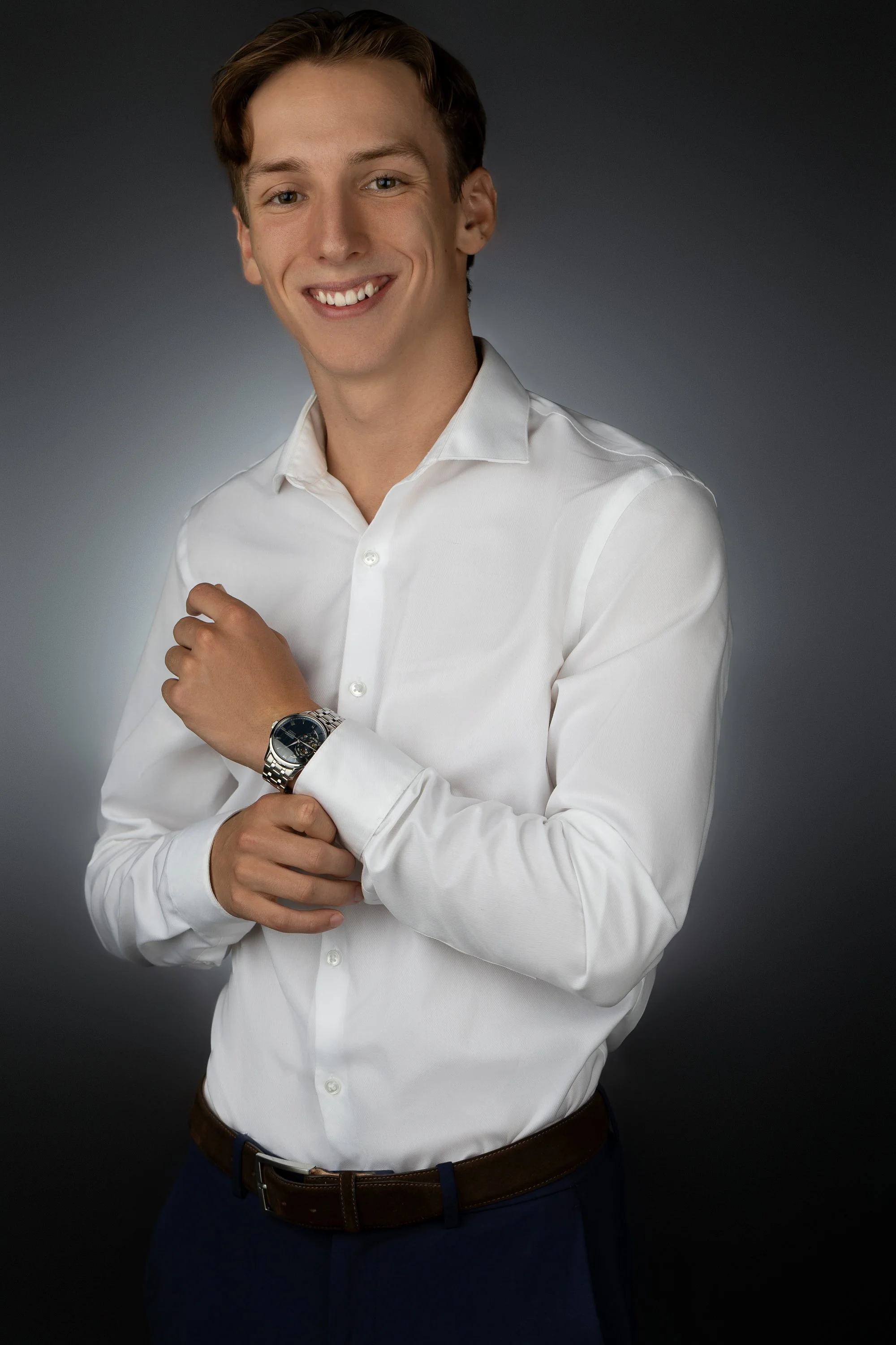 Portrait of a young man with light skin, wearing a white dress shirt, dark pants, and a wristwatch, smiling and posing with one hand on his chest against a dark gradient background.