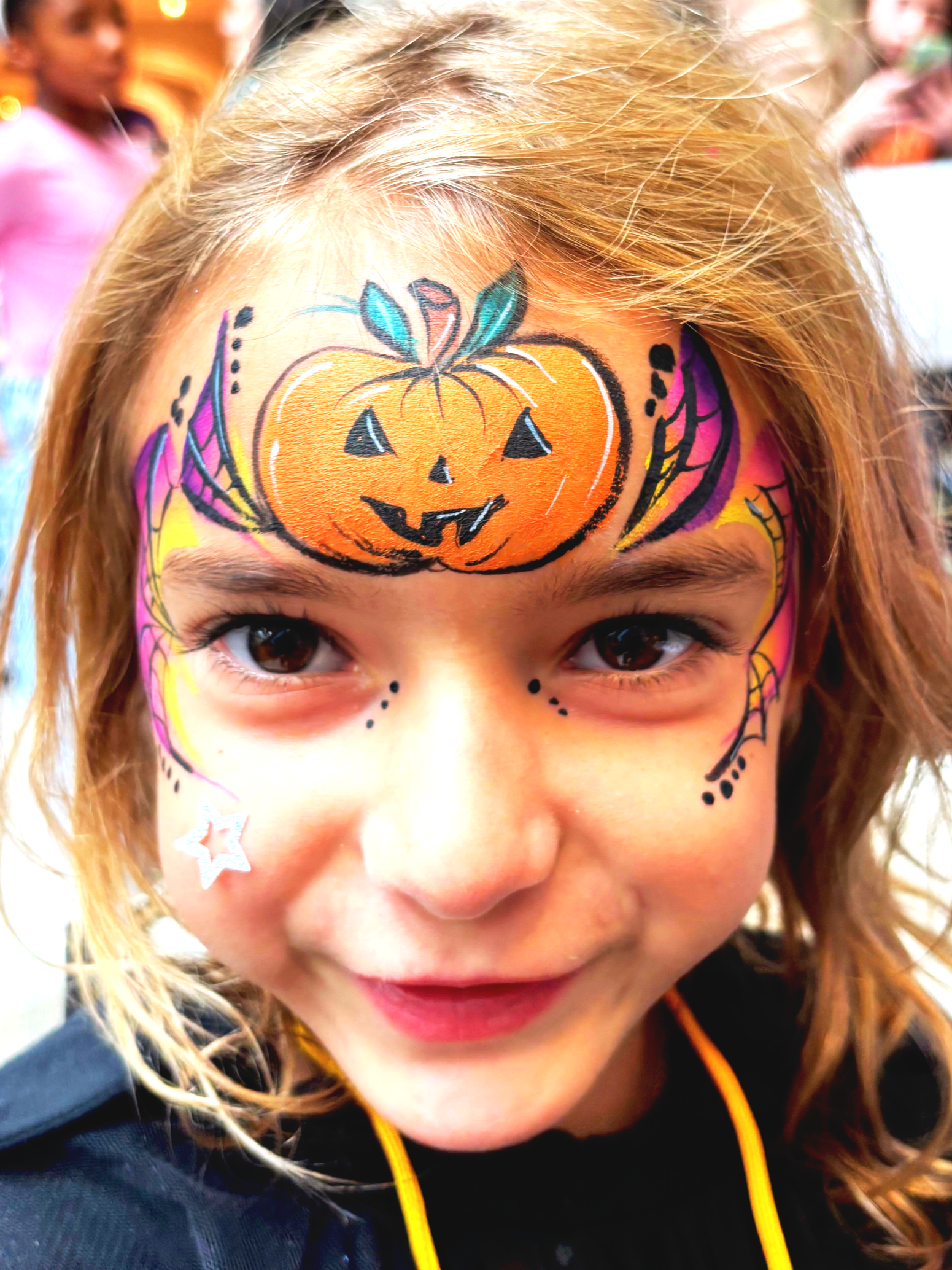 Close-up of a young girl with face paint of a pumpkin on her forehead, various colorful details, and a star sticker near her mouth, smiling with brown eyes and light brown hair.