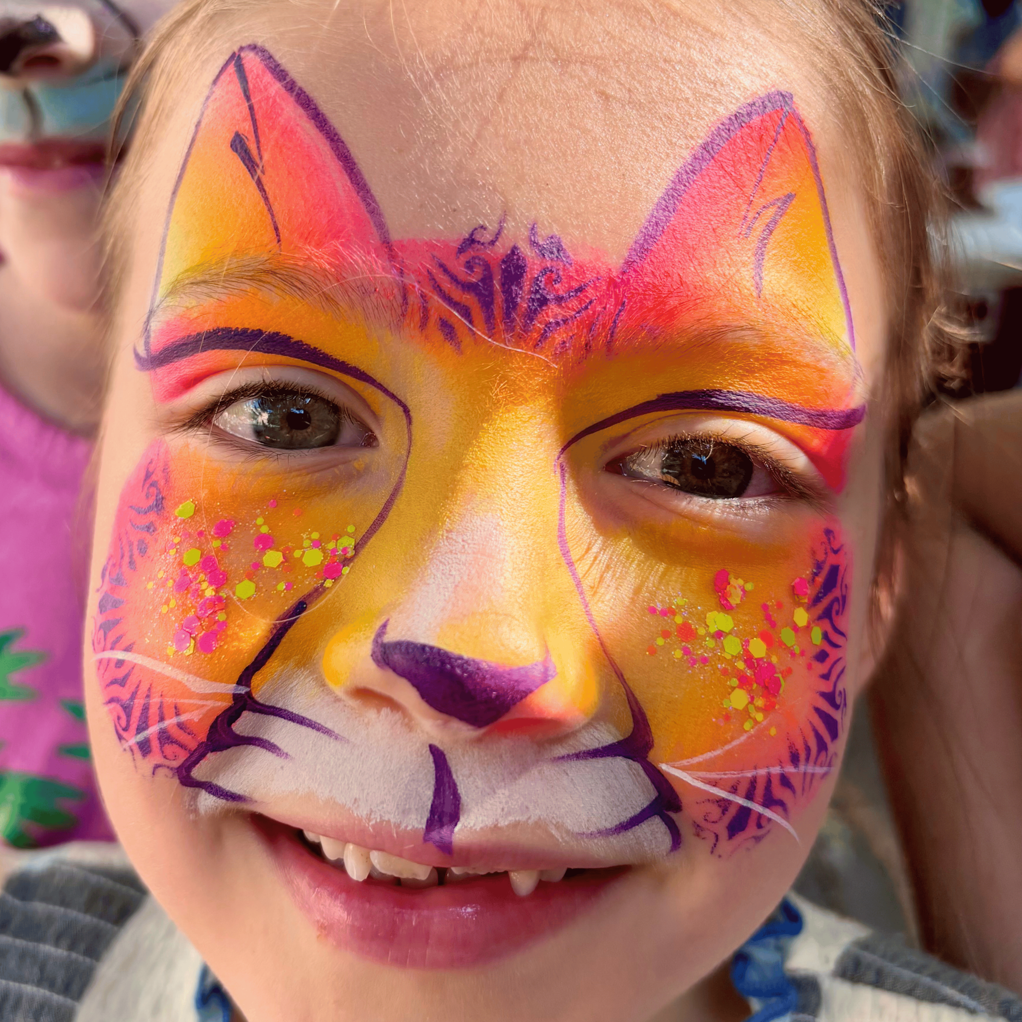 Close-up of a child's face with colorful kitty face paint, including purple, orange, and pink hues, and glitter with pink and yellow spots on the cheeks.